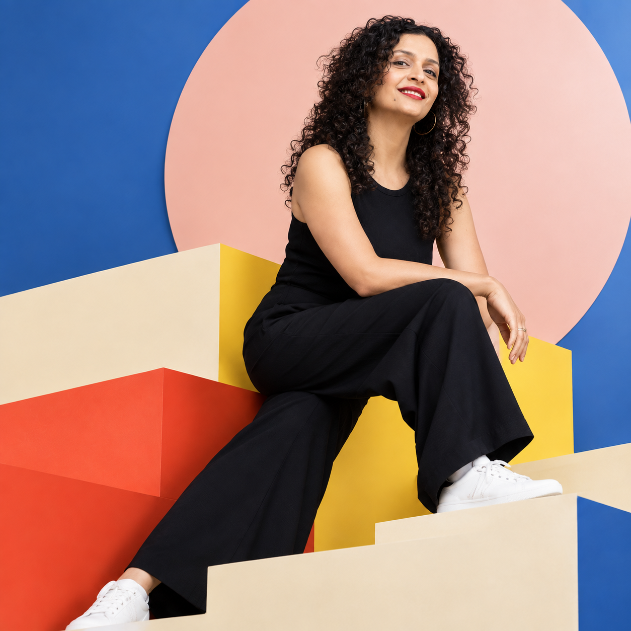 A woman with curly dark hair wearing a black sleeveless top, black pants, and white sneakers sitting on colorful geometric steps with a pink circle and blue background.