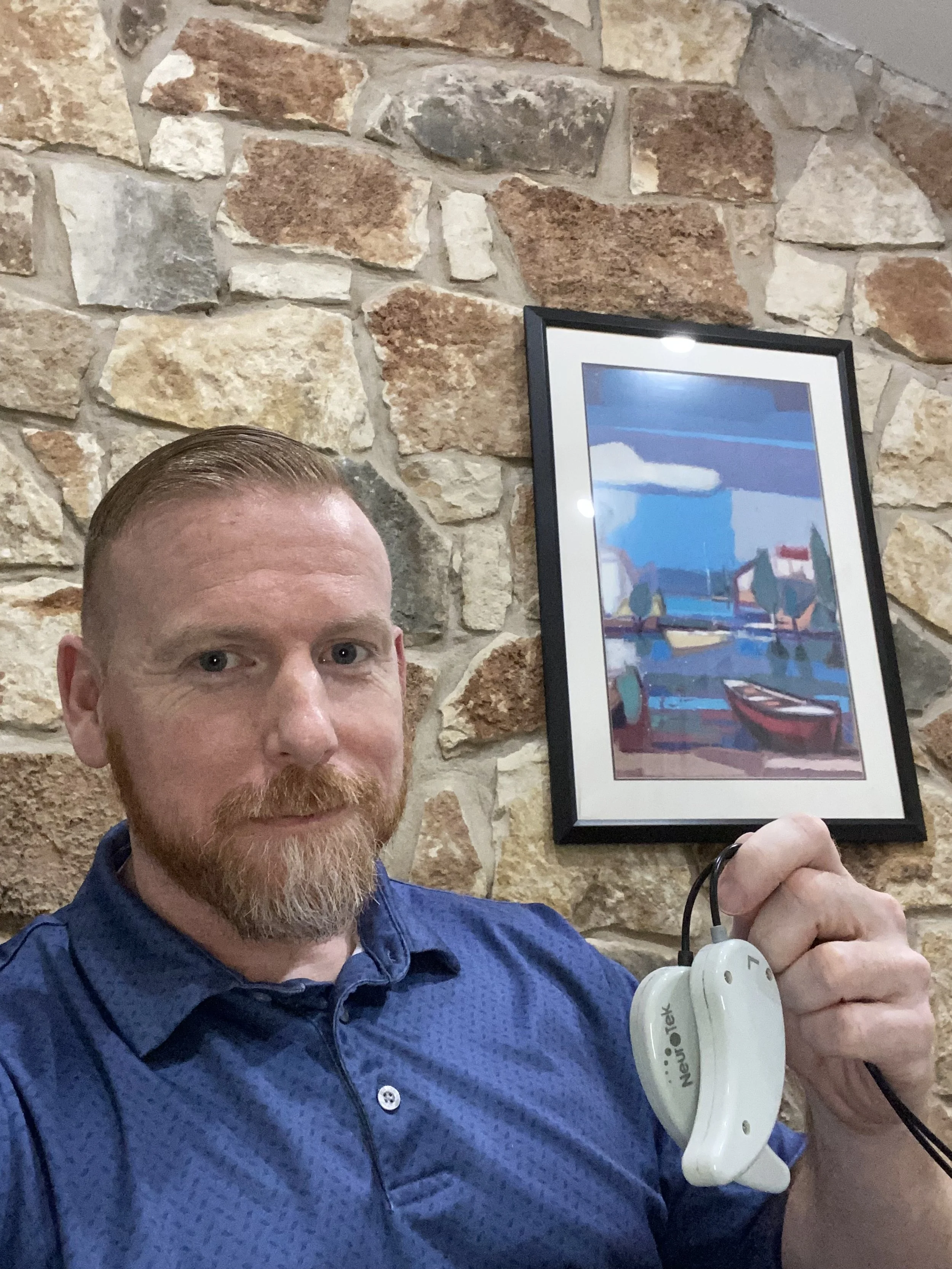 Founder Alex Penrod in front of rock wall with picture hanging in background holding up EMDR tappers used for bilateral stimulation.