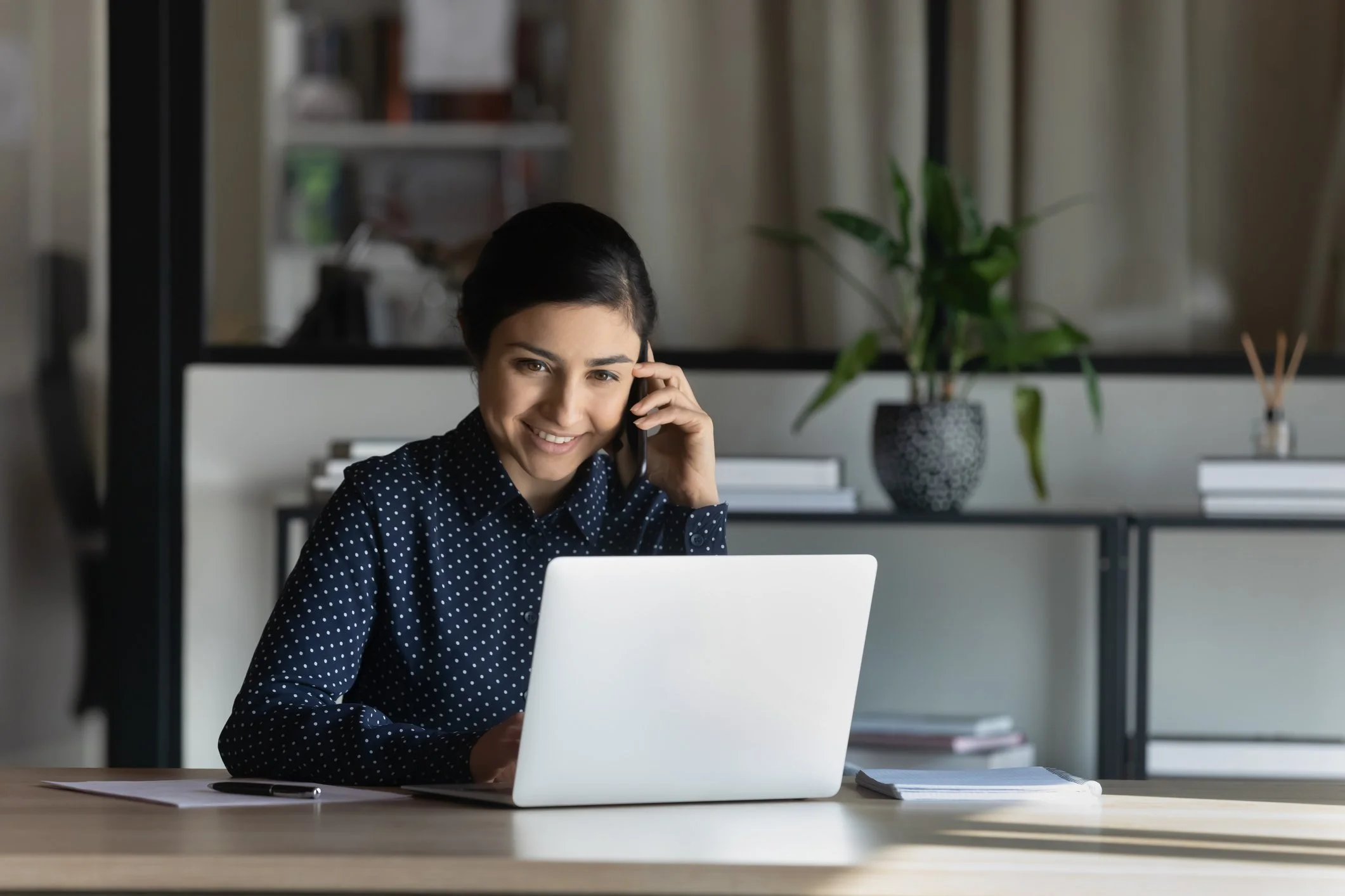 A woman with dark hair, wearing a navy blue polka dot blouse, smiles while talking on a mobile phone and booking a free 15-minute consultation with Neuro Nuance Therapy and EMDR, PLLC.
