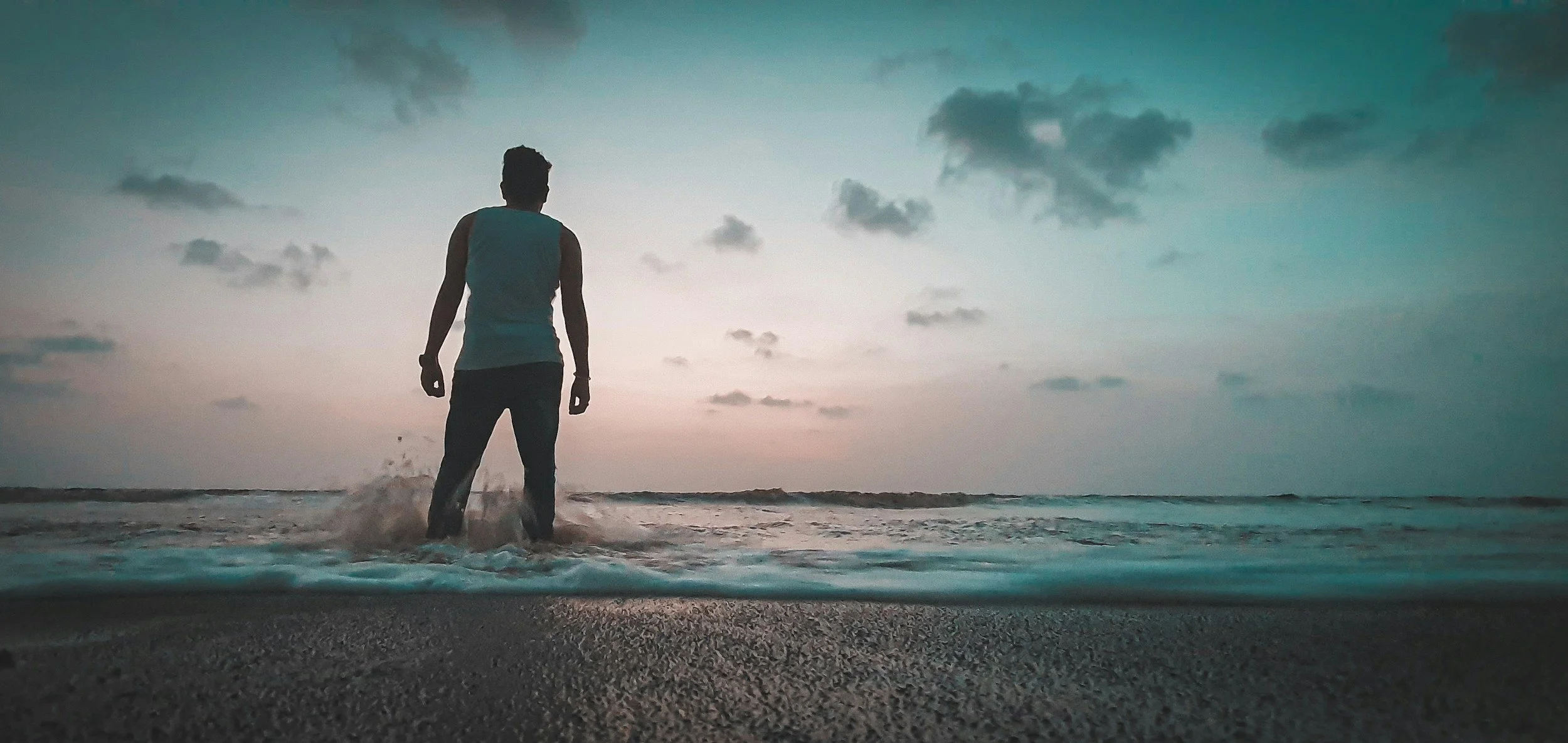 A person standing in the ocean at sunset or sunrise, looking out at the water and sky.