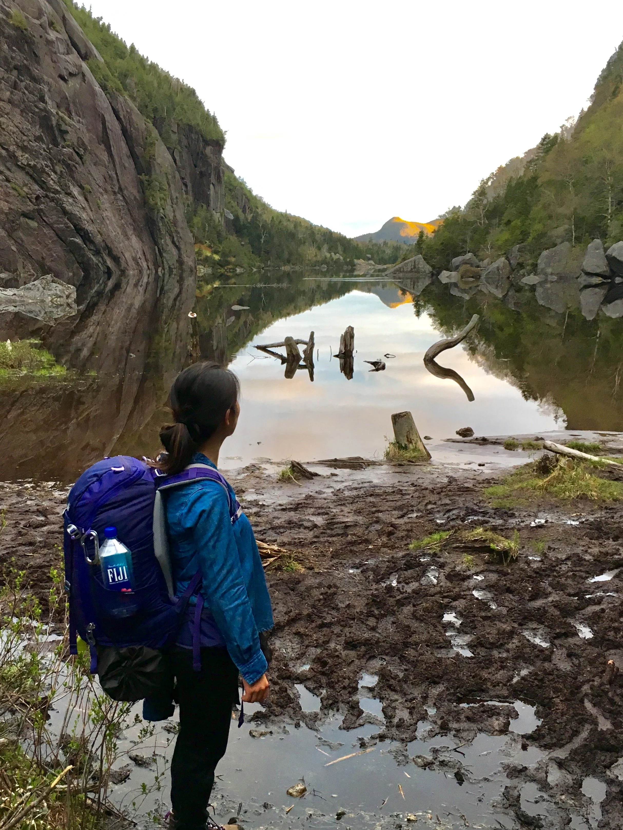 Hiker standing on muddy trail beside a calm river reflected between forested mountains, with some driftwood and rocks in the water, and a mountain in the distance.