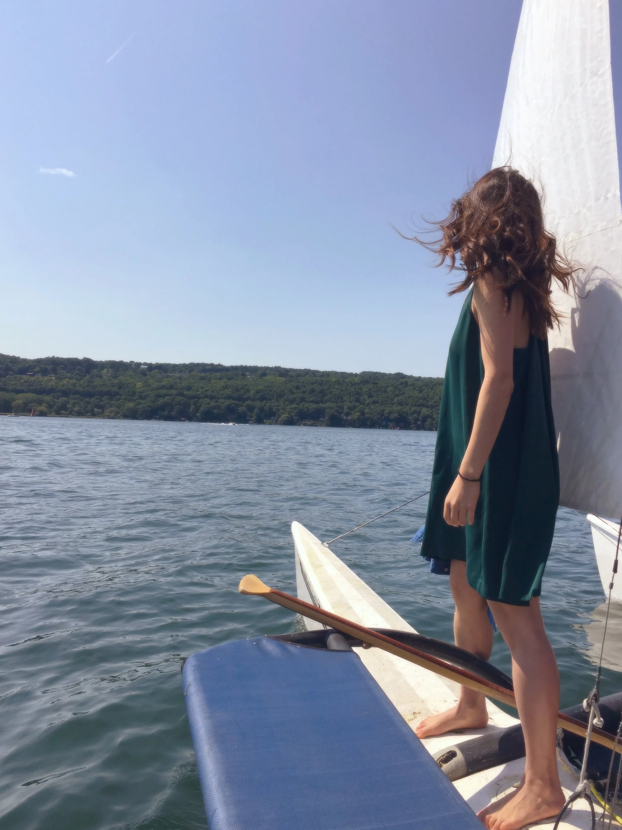 A young woman in a teal dress standing on a sailboat near the edge, with her hair blowing in the wind, looking out towards a lake and distant green hills under a clear blue sky.