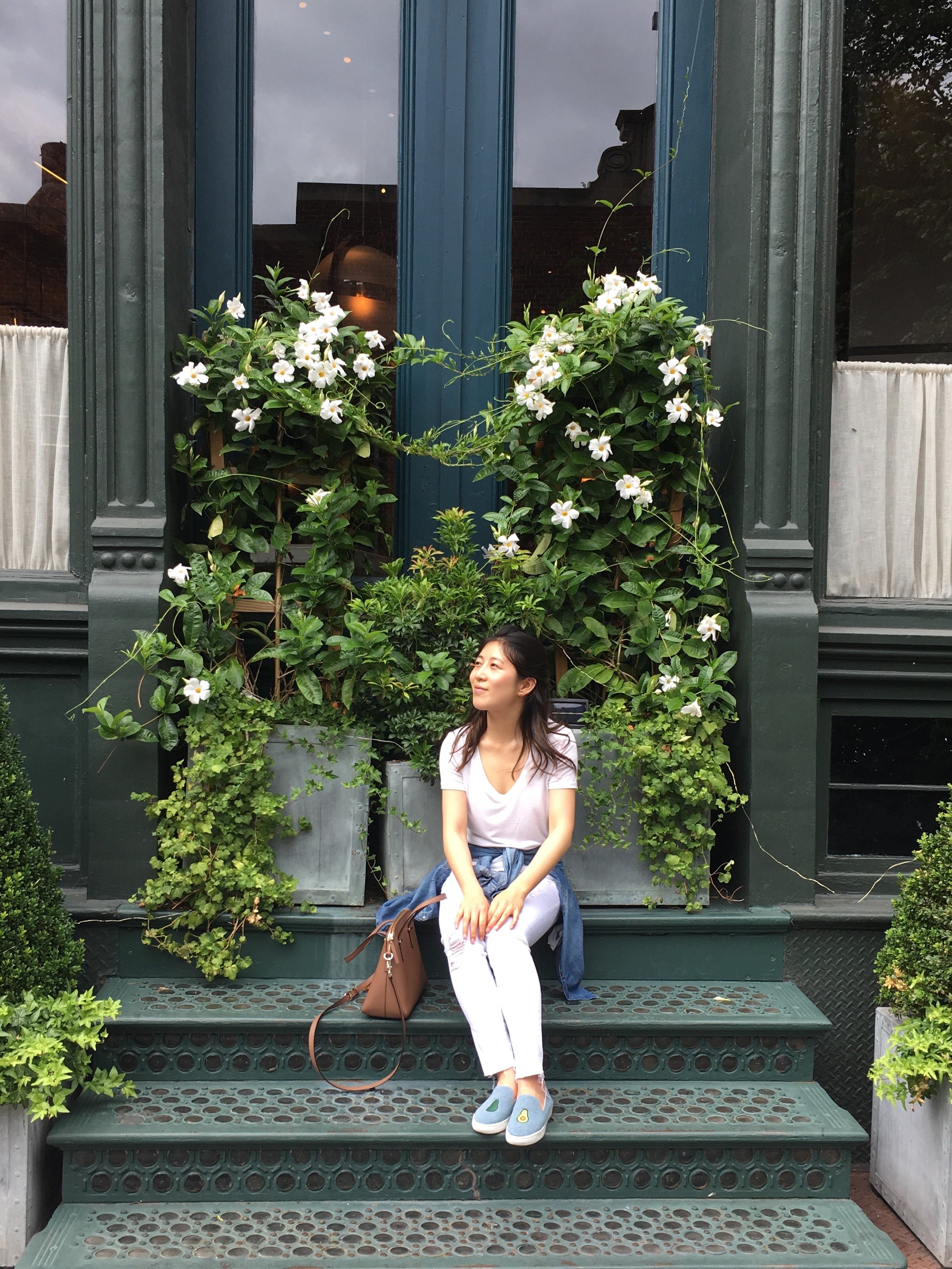 A woman sitting on metal steps in front of a building with large windows and green plants with white flowers.