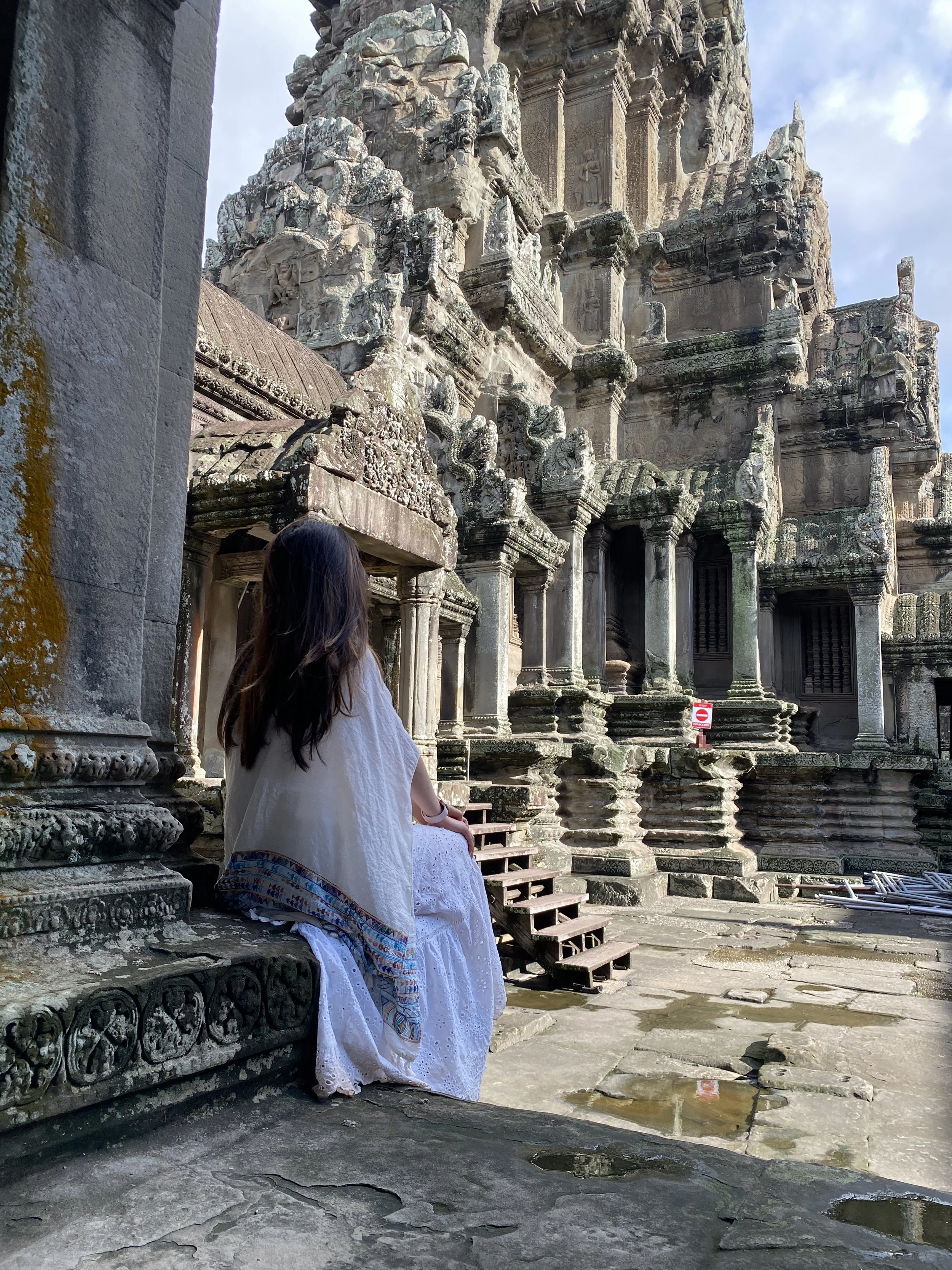 A woman in a white dress sitting on a stone ledge, looking at an ancient stone temple with intricate carvings, moss, and lichen growths.