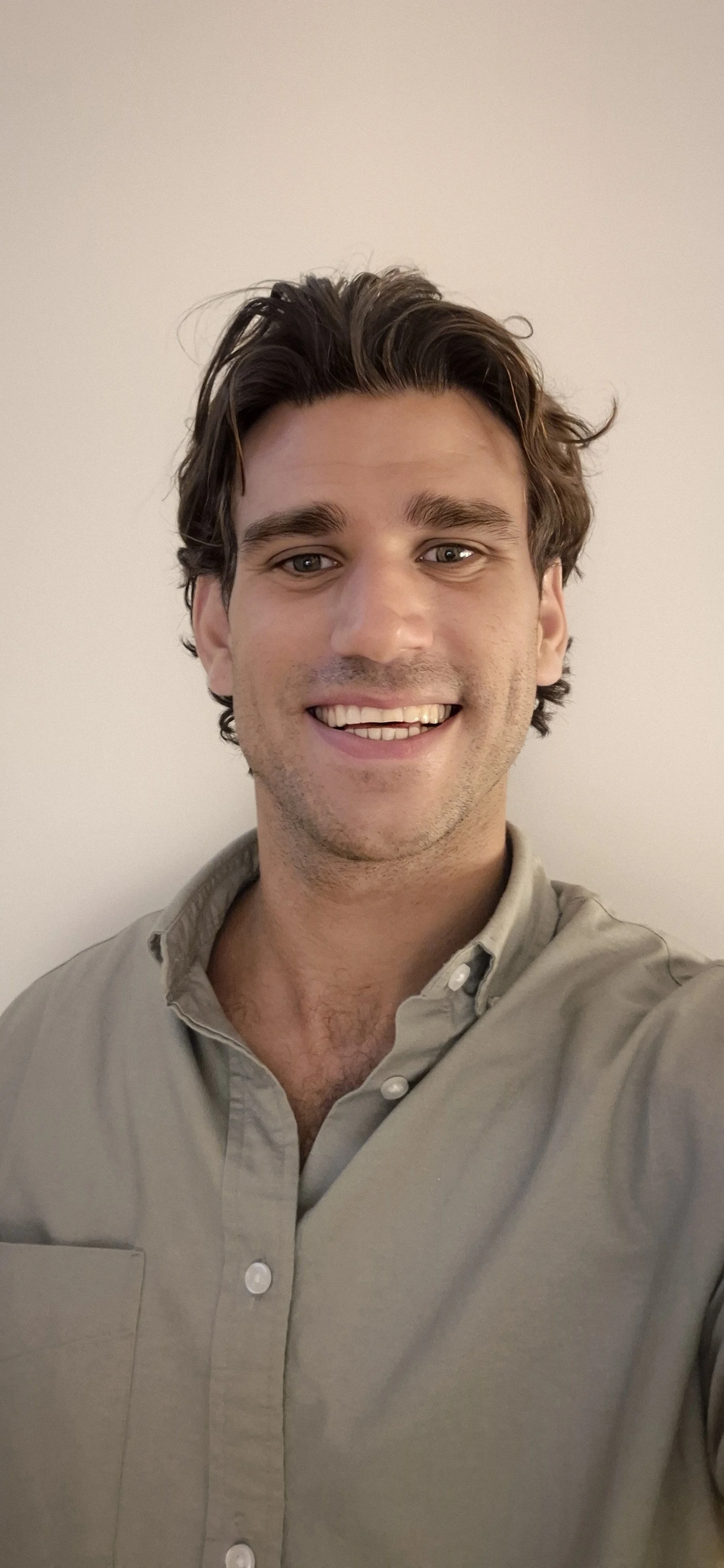 Close-up of a smiling young man with wavy brown hair, a small earring, and a black shirt against a gray background.
