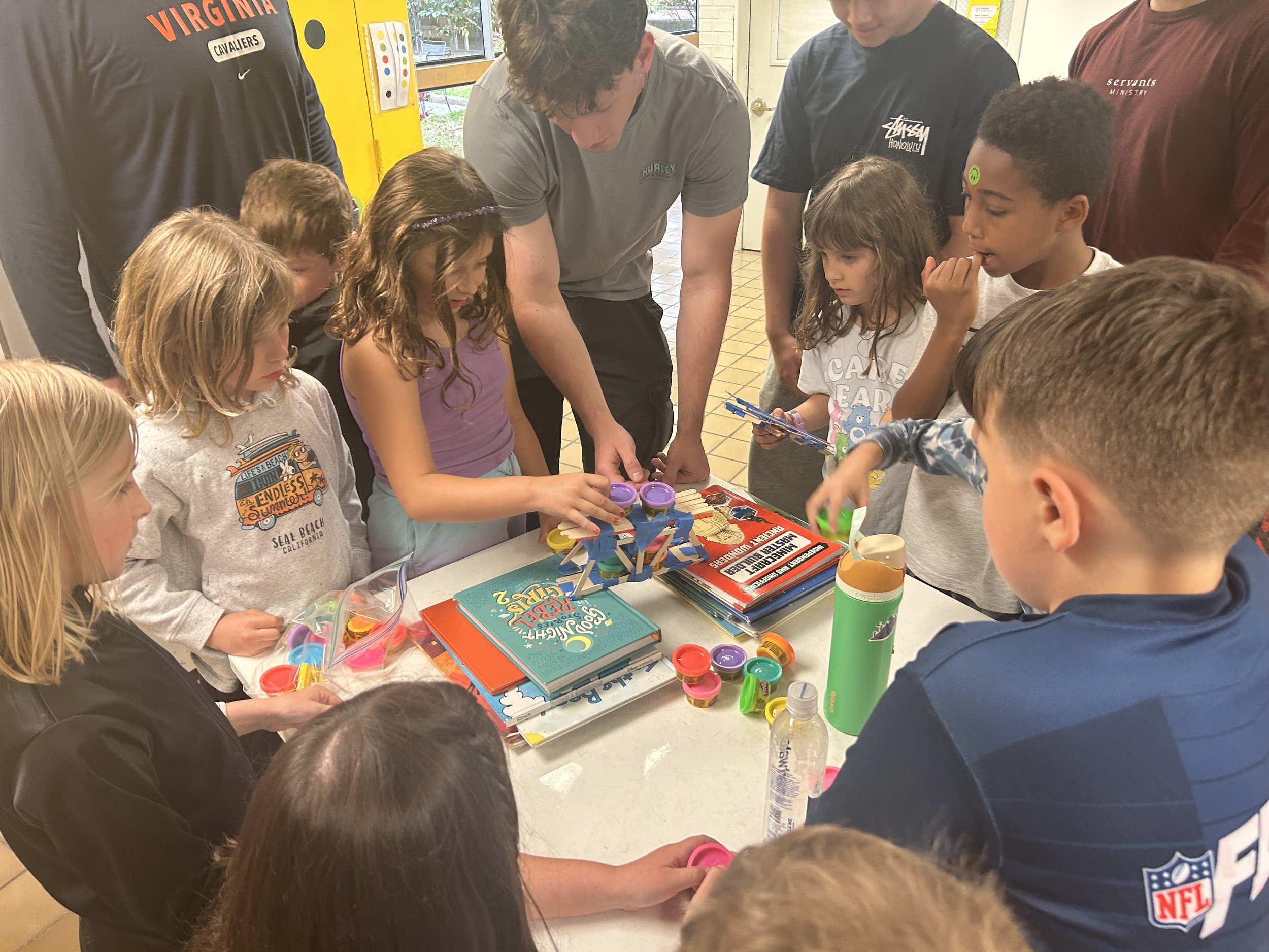 A group of children and young adults gathered around a table playing a game with colorful plastic parts and toys in a bright indoor space.