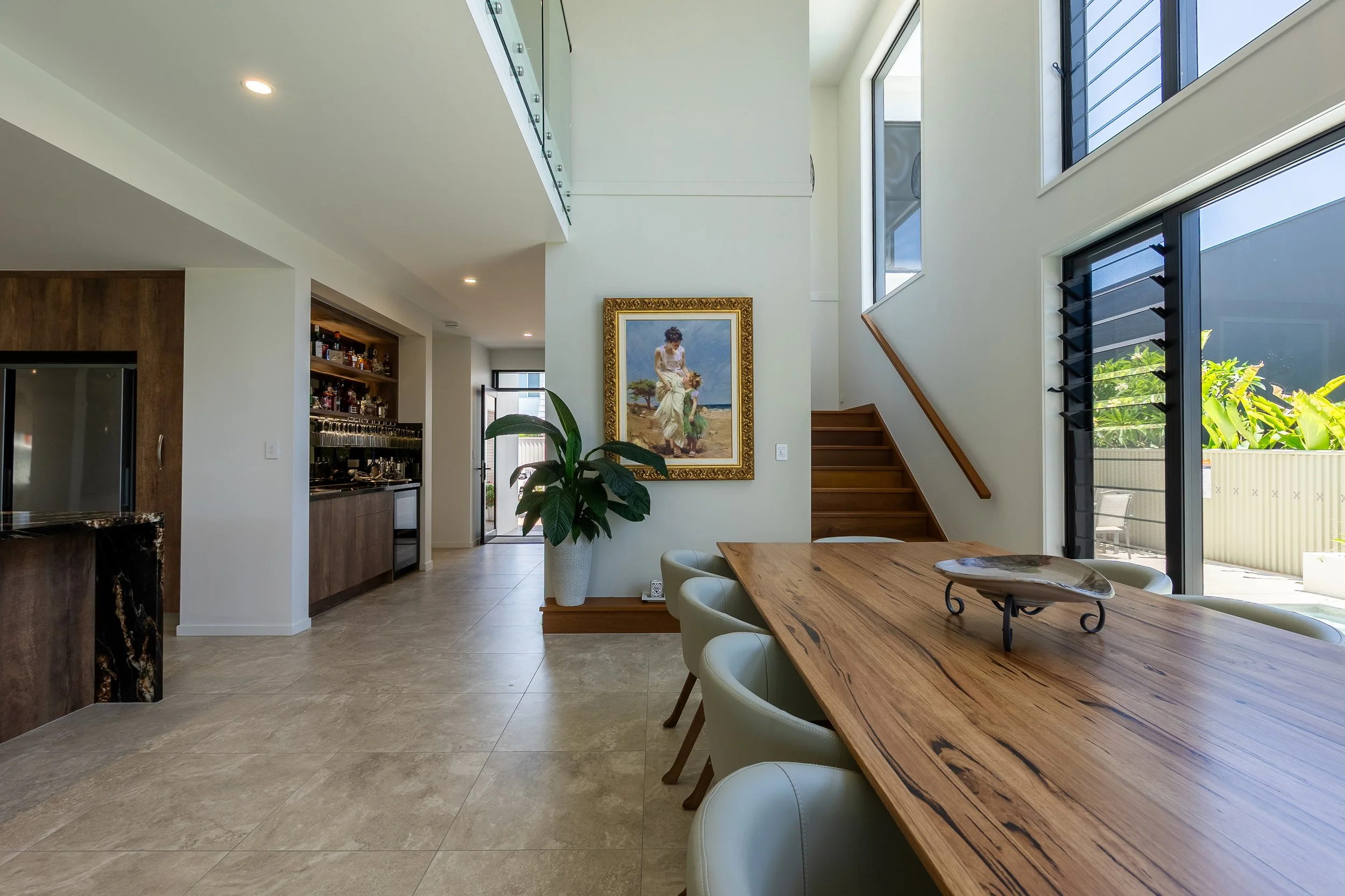 Interior view of a modern home with high ceilings, large windows, a wooden staircase, a framed painting of a woman with a child outdoors, a large potted plant, a wooden dining table with white chairs, a bar area with glassware, and outdoor patio visible through sliding glass doors.