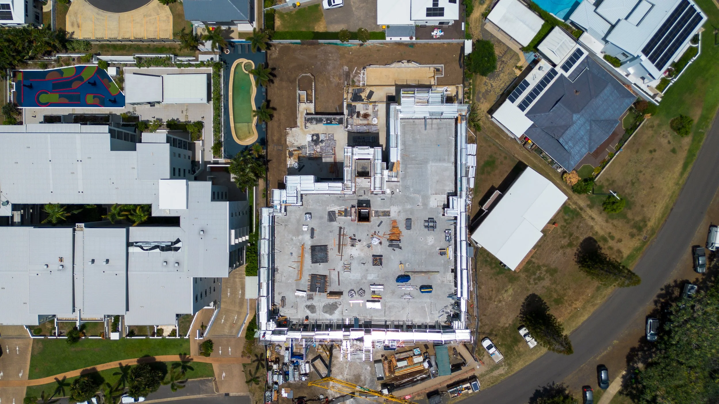 An aerial view of a building under construction with a white roof, surrounded by other completed buildings, a playground, trees, parked cars, and nearby roads.