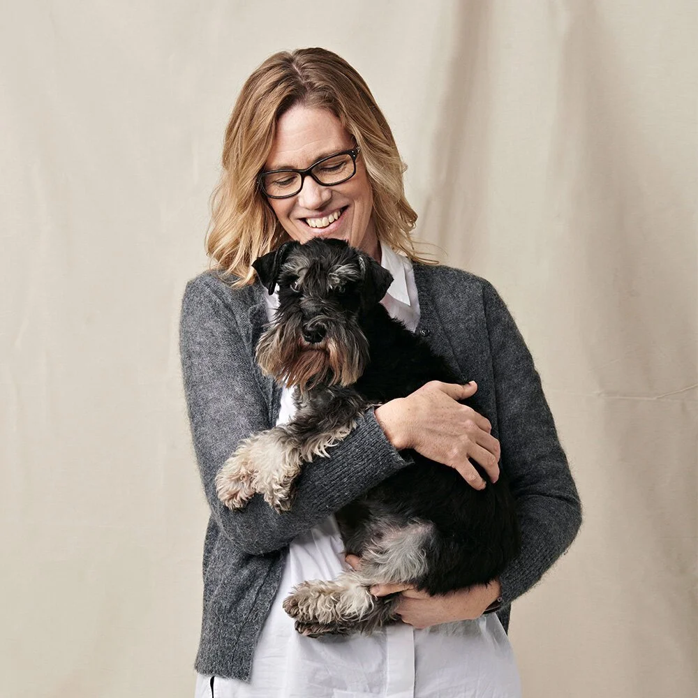 Kvinne med brunt, skulderlangt hår og briller smiler og holder en liten svart og grå hund.