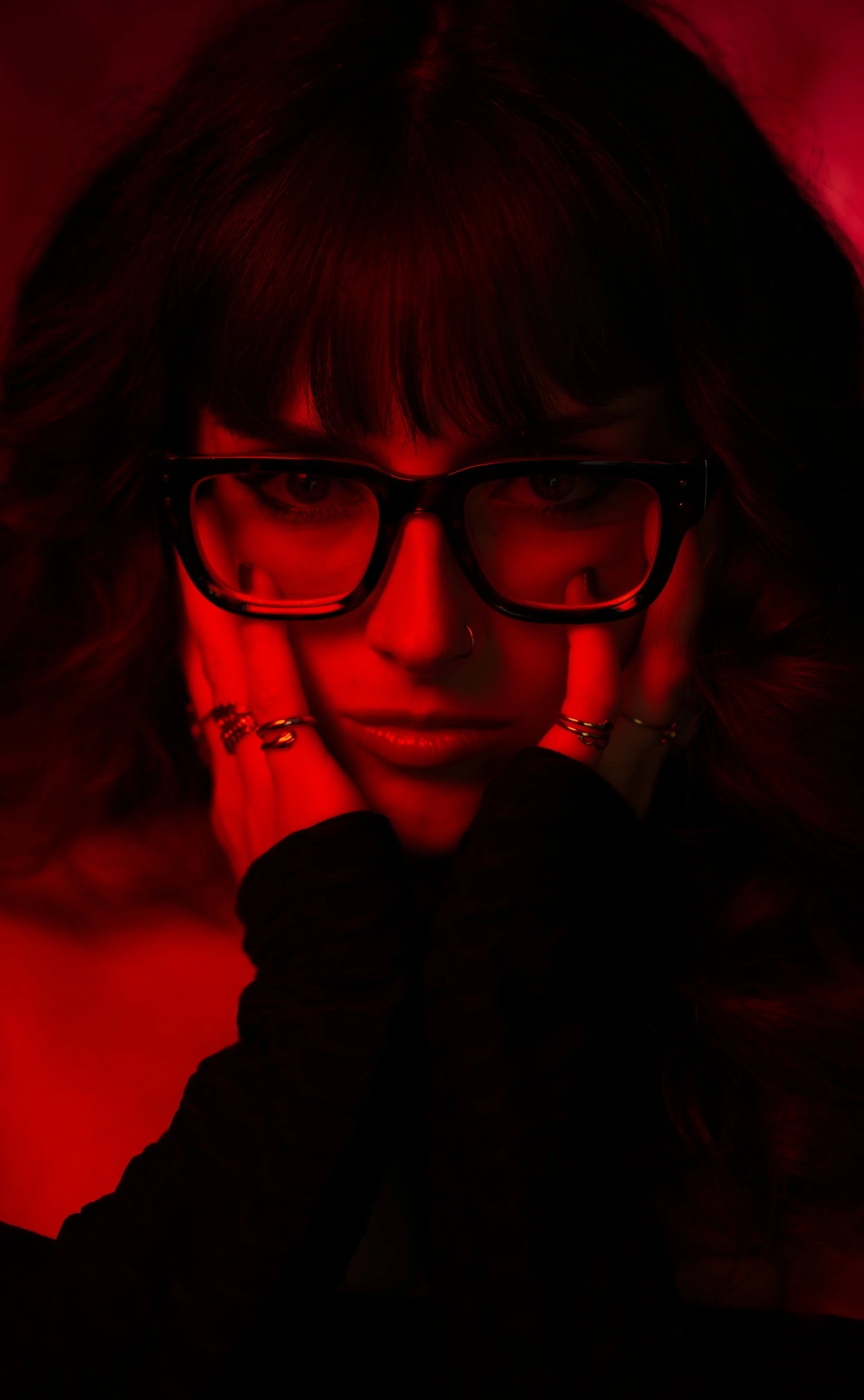 A woman with dark hair and glasses, holding her face with her hands, illuminated in red light.