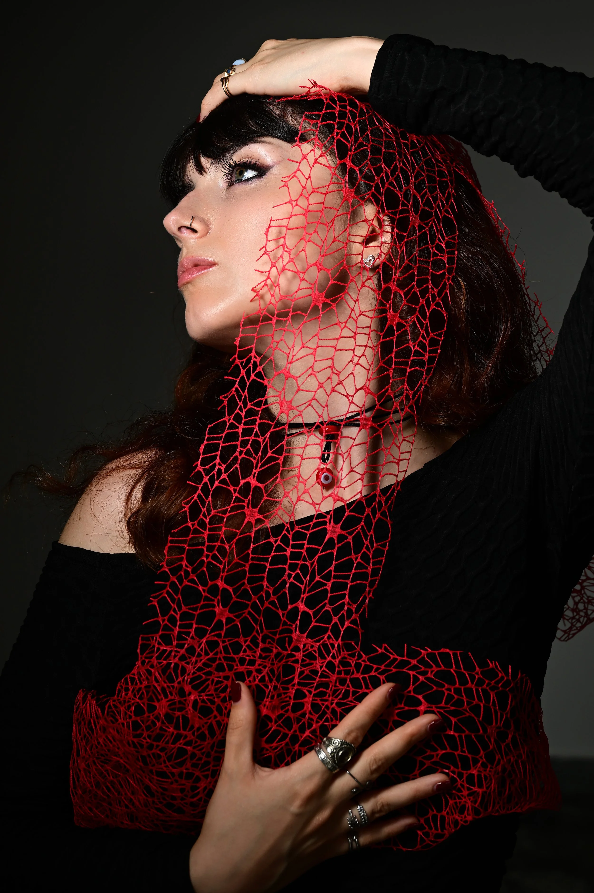 A woman with dark hair and makeup holding a red lace veil, wearing silver rings, in a black long-sleeve top.