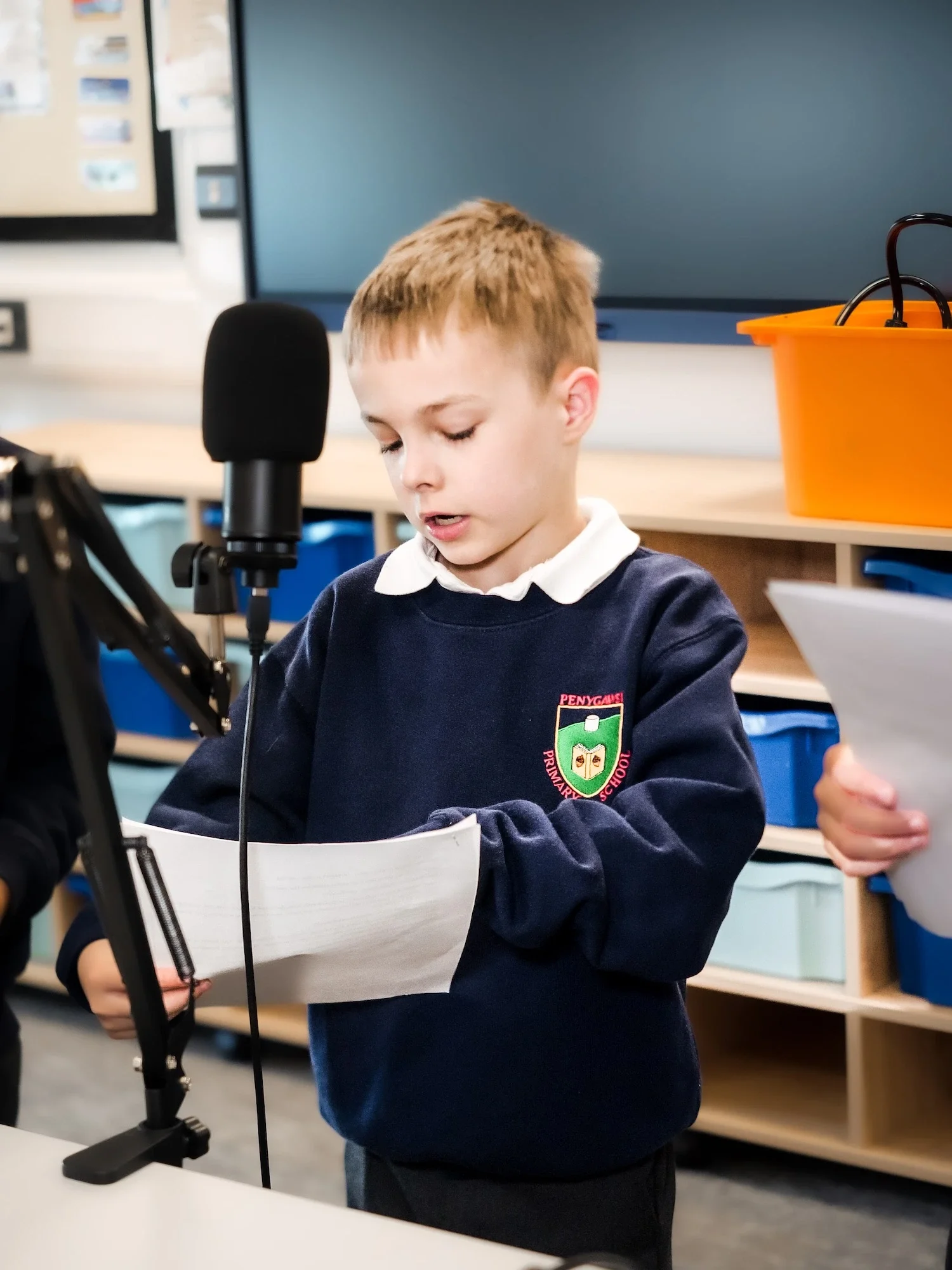Young boy in school uniform speaking into a microphone while reading from a paper in classroom.