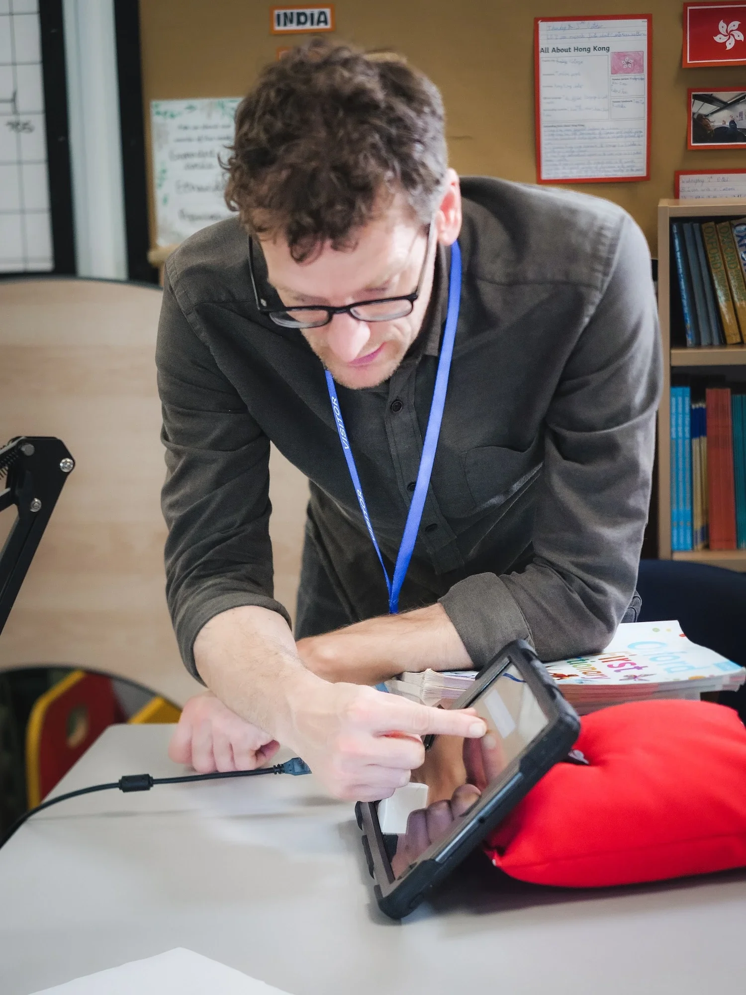 A male teacher with glasses wearing a gray shirt and a blue lanyard, leaning over a table, using a tablet.