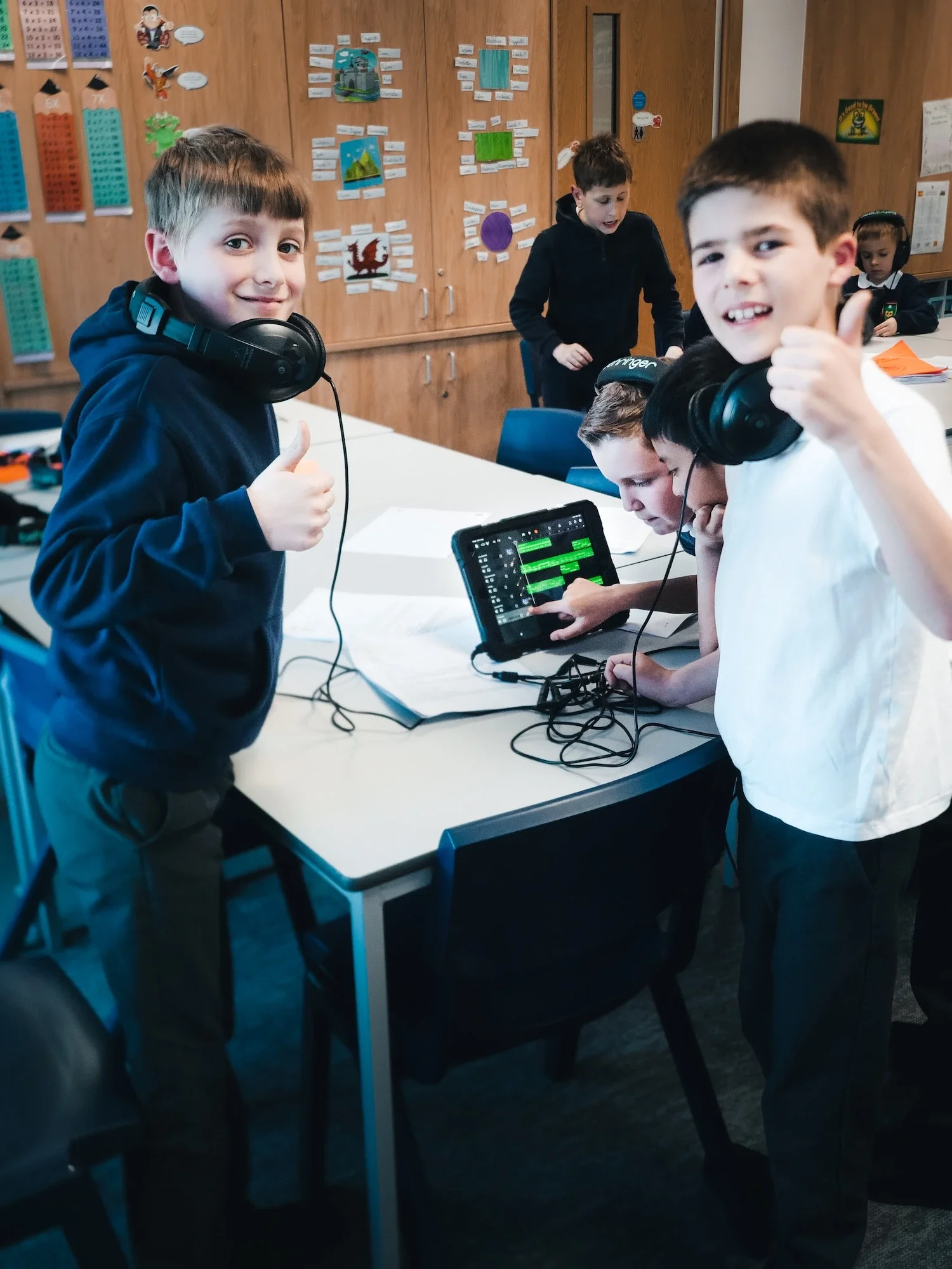 Two boys in a classroom wearing headphones, giving thumbs up, with a tablet and music editing software on the table, and other children working and engaging in activities in the background.