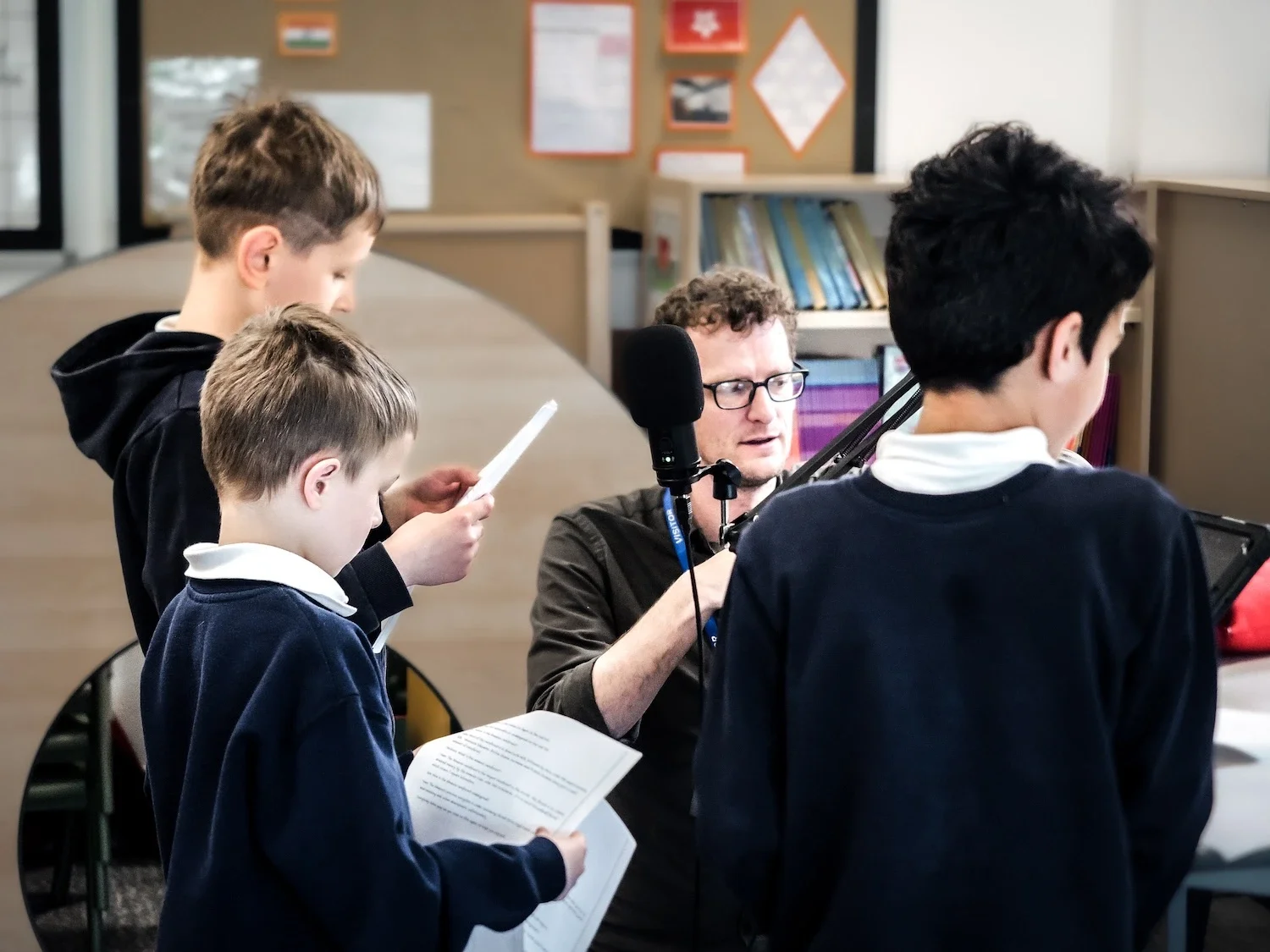 Four students, three boys, wearing school uniforms, stand around a man with glasses, recording a podcast or recording session with a microphone in a classroom.