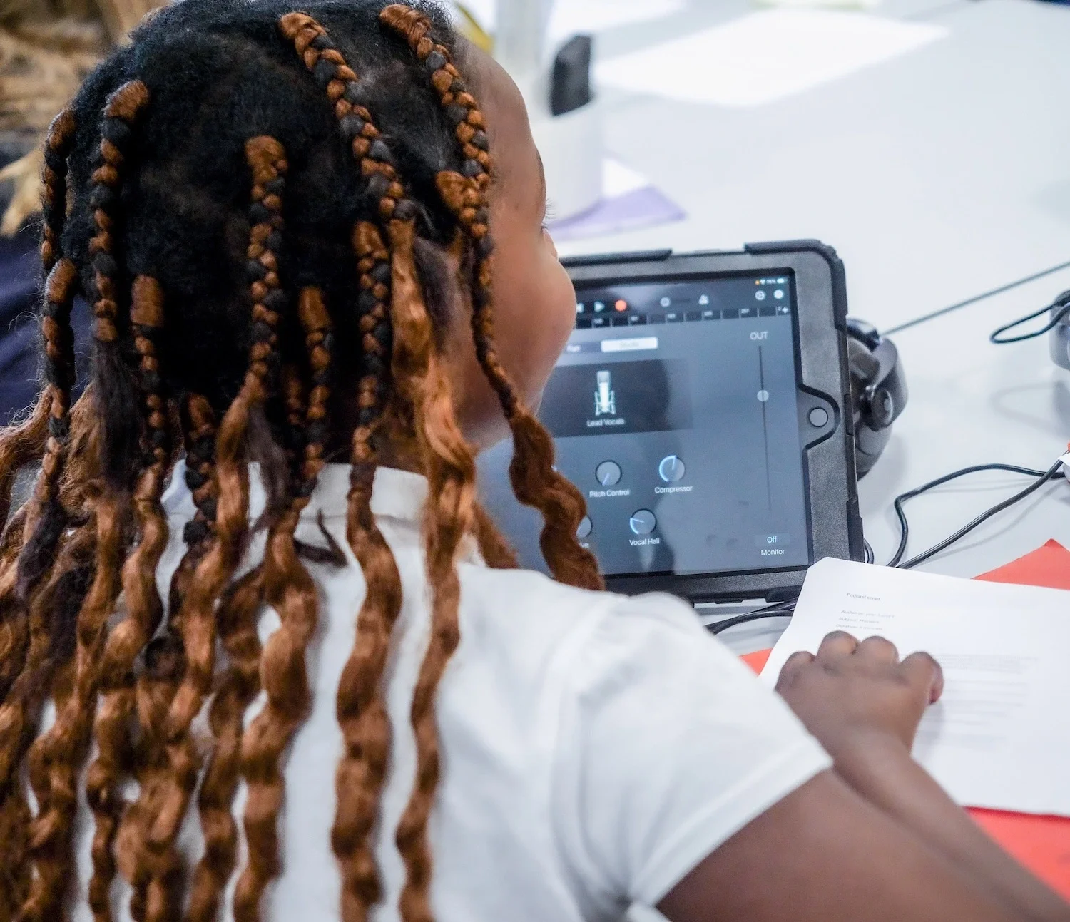 Person with braided hair looking at a tablet with music production software, sitting at a white table with a notebook nearby.