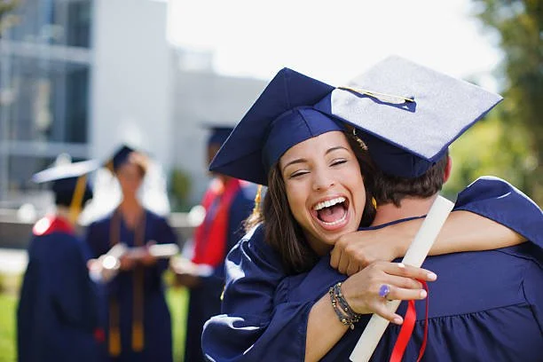 Smiling young woman in graduation gown and cap holding diploma with red ribbon, celebrating outdoors with other graduates in the background.