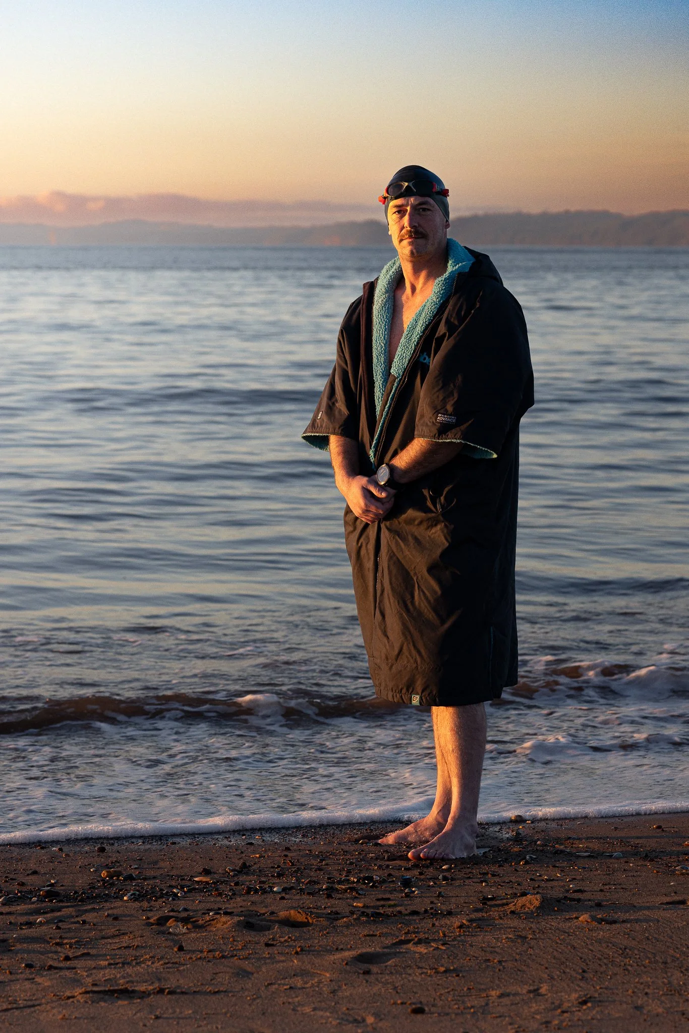 A man, dressed in a dry robe, swimming cap and goggles stands at the edge of the sea, on a sandy beach. The sky behind him is golden and clear.