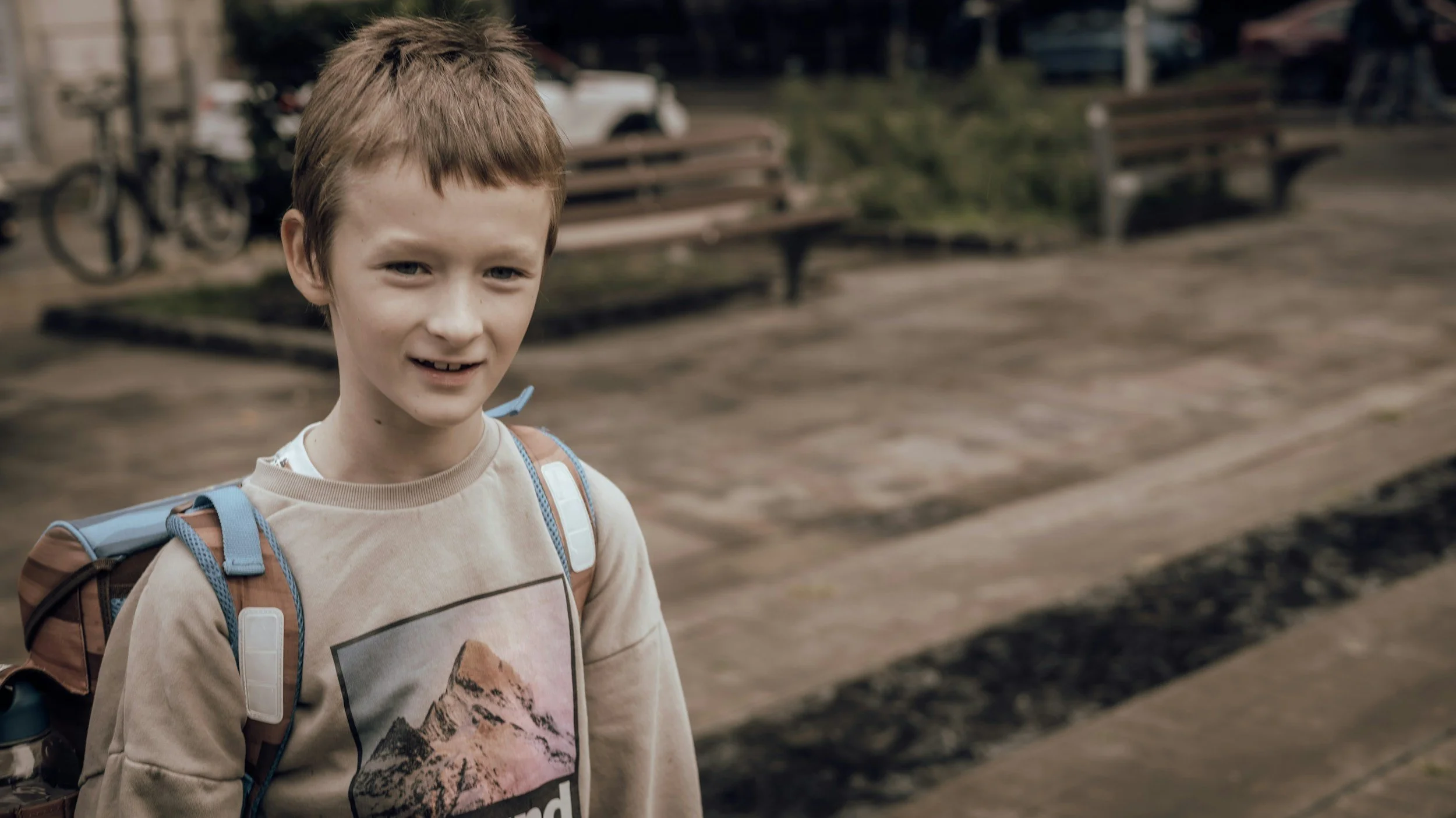 A young boy with red hair and a beige sweatshirt standing outdoors with a backpack, smiling slightly, with benches and parked bicycles visible in the background.