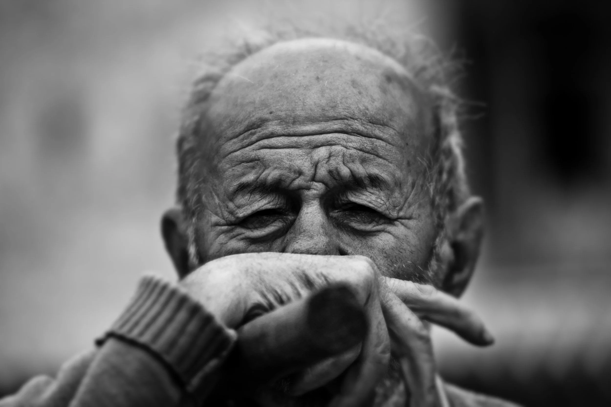 A close-up black and white photo of an elderly man with wrinkles, covering his mouth with his hand, wearing a striped sweater.