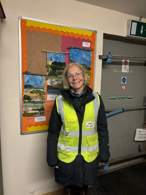 A smiling woman wearing glasses and a bright yellow safety vest standing indoors in front of a colorful bulletin board with landscape artwork and a door with safety signs.