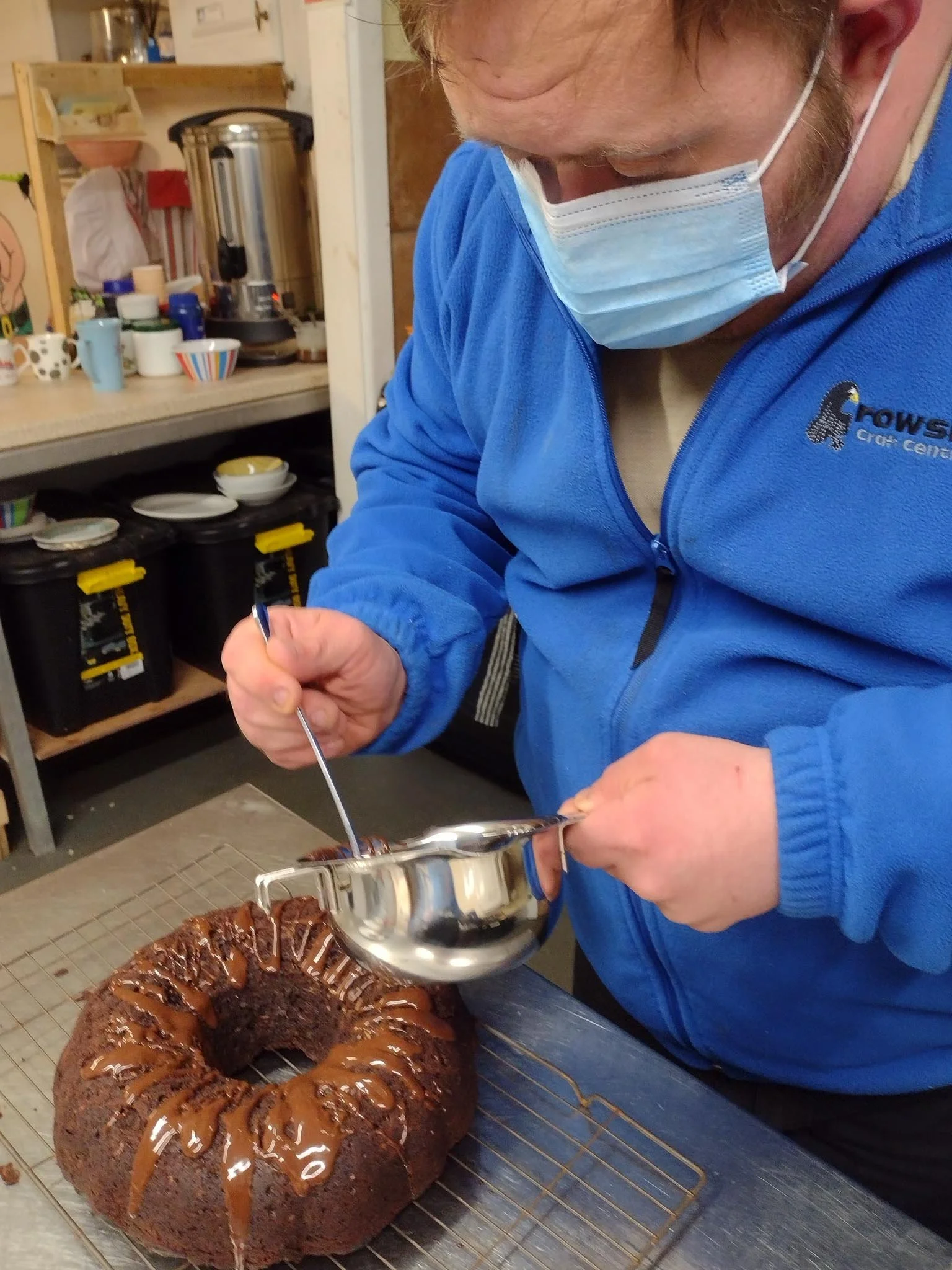 A man wearing a blue fleece jacket and a medical mask is pouring chocolate sauce over a bundt cake on a cooling rack in a kitchen.