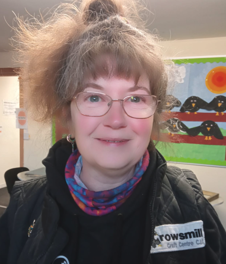 A woman with curly, frizzy hair pulled up in a ponytail, wearing glasses, a colorful scarf, and a black jacket with a Trowmill Craft Centre badge, smiling in a room with a colorful, animal-themed bulletin board in the background.