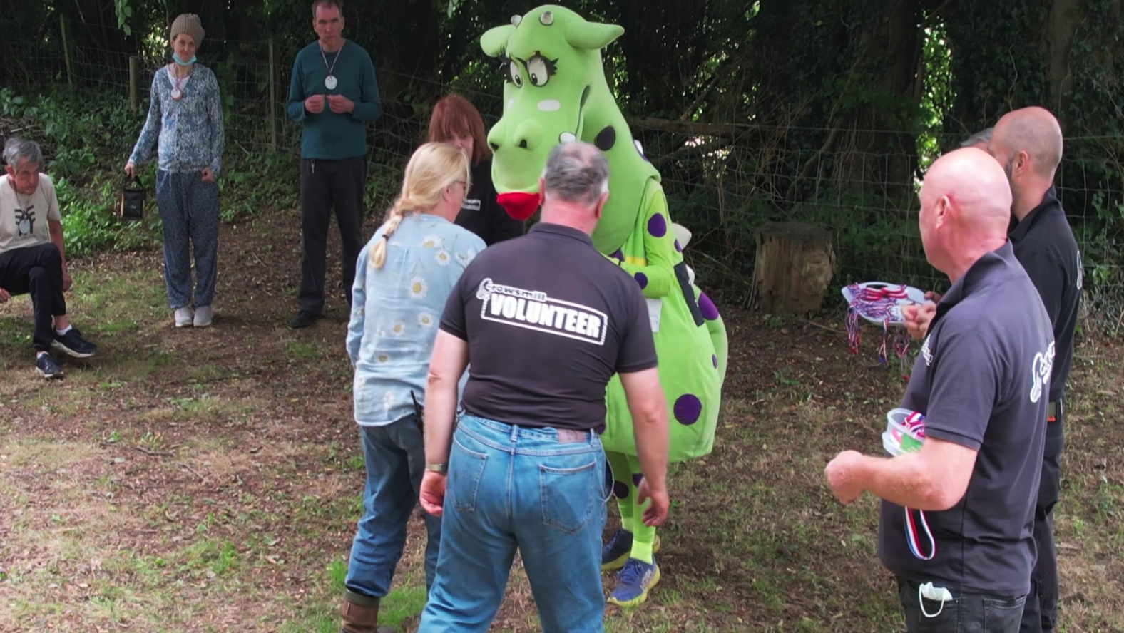 People gathered outdoors around a person in a green and purple cow mascot costume, with some wearing volunteer shirts, during an event in a wooded area.