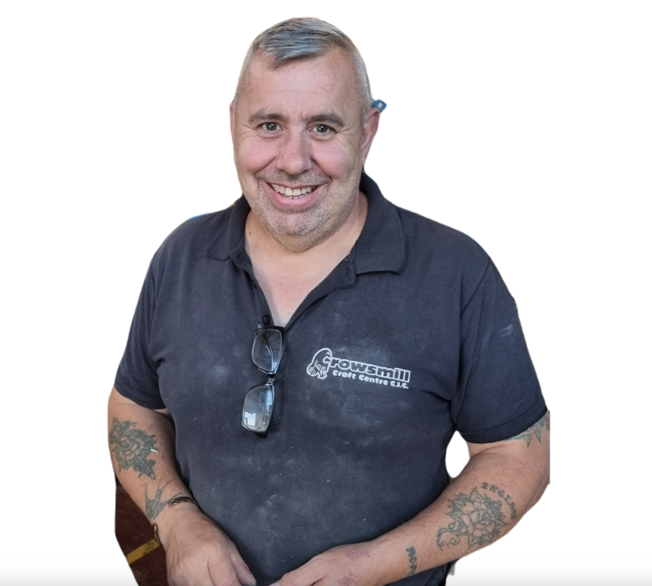 A middle-aged man with short gray hair and a beard, smiling, wearing a black polo shirt with a logo that says 'Fowsmil Craft Centre' and glasses hanging from the collar. Tattoos are visible on his arms.