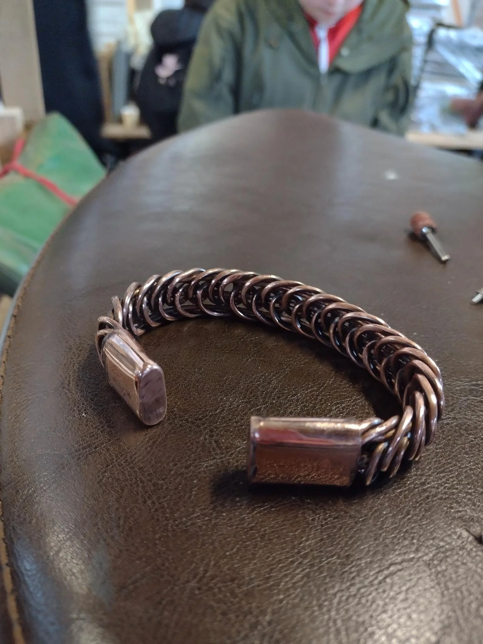 A bracelet made of interlinked metal rings with rectangular metal end caps, resting on a dark brown leather surface.
