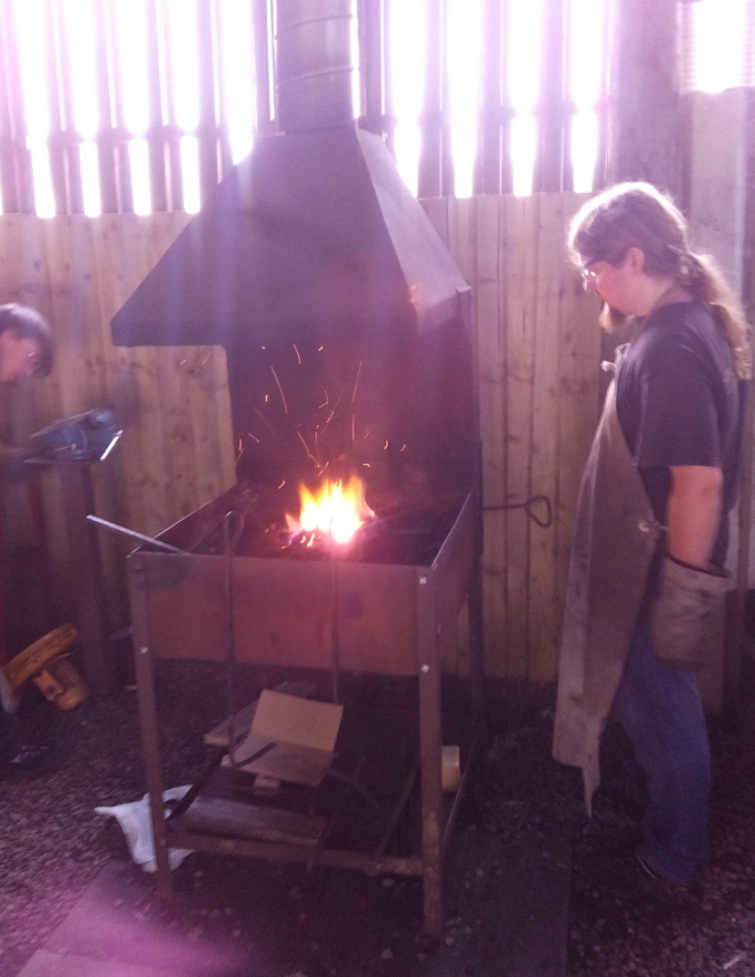 Person wearing a black shirt and apron standing next to a forge with a fire inside, sparks flying, in a workshop with wooden walls and tools.