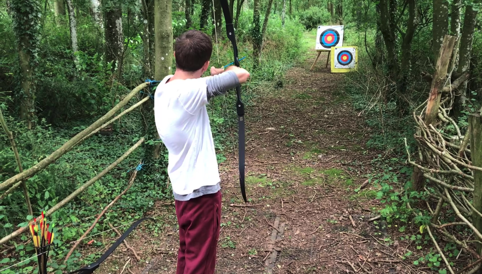 A young boy in a forest shooting an arrow with a bow at a target in the distance.