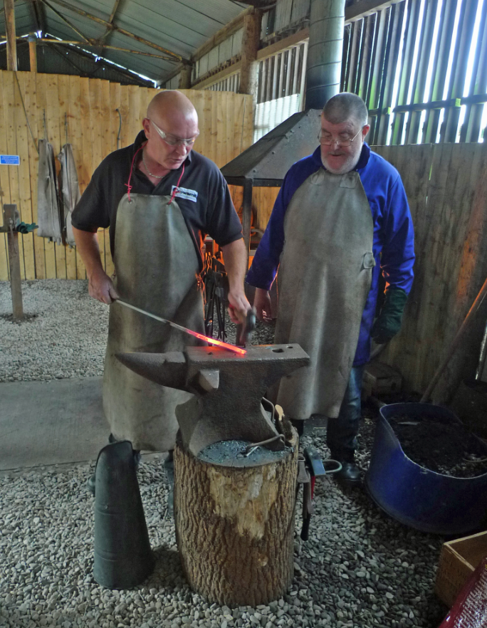 Two men forge metal outdoors, wearing aprons and safety glasses, with one heating metal in a forge and striking it on an anvil set on a tree stump.