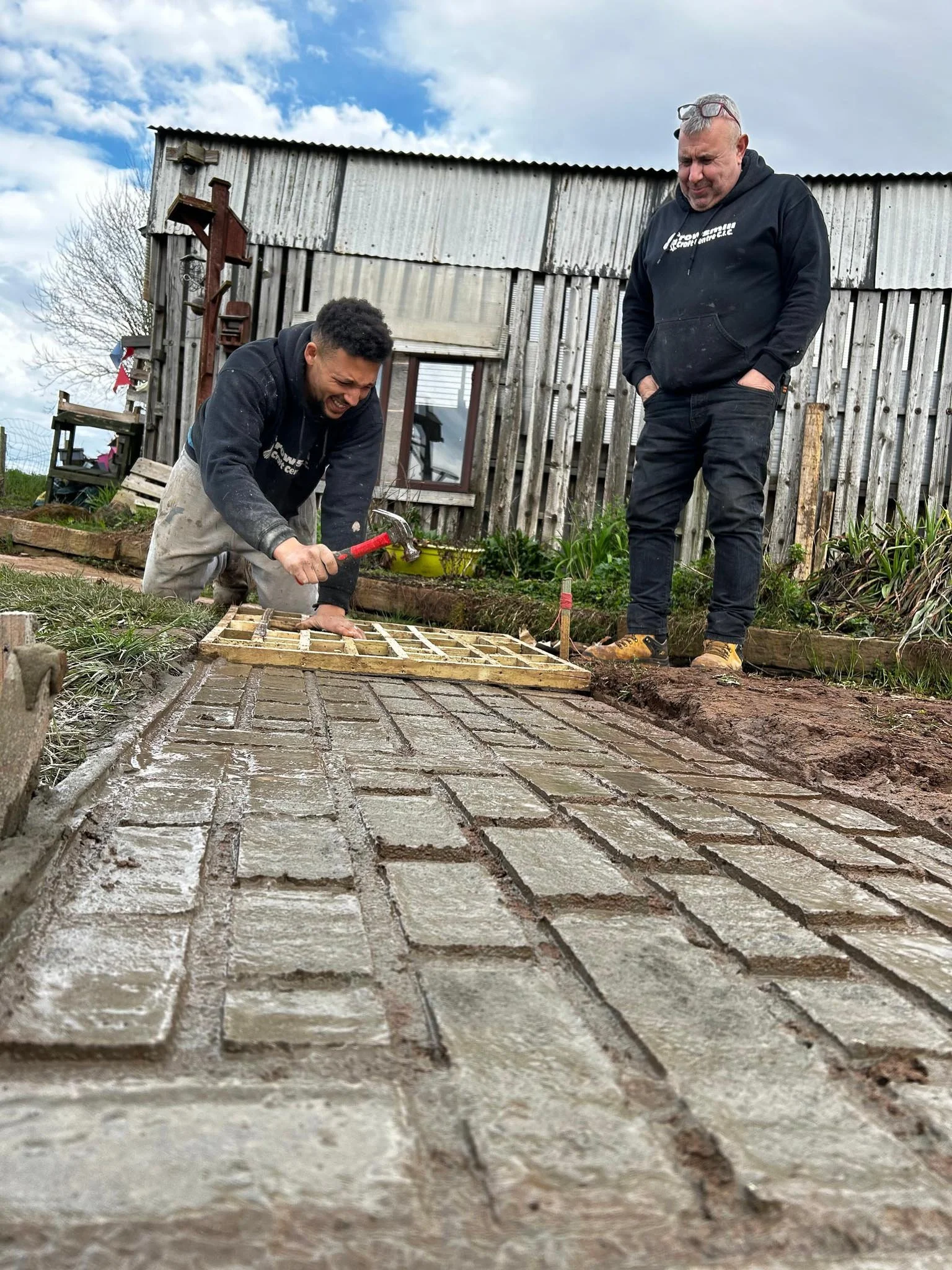 Two men laying bricks on a pathway outdoors, one is working with a hammer while the other watches, with a weathered wooden shed in the background and a cloudy sky overhead.