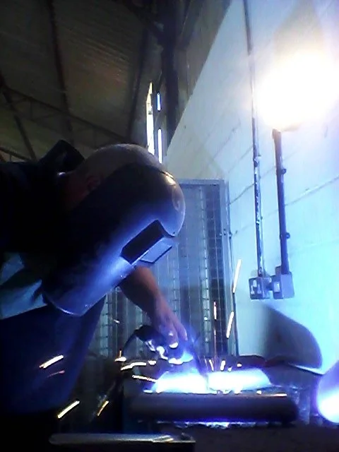A person welding metal in an industrial workshop, wearing a welding helmet and protective gear, with bright sparks and light from the welding process.