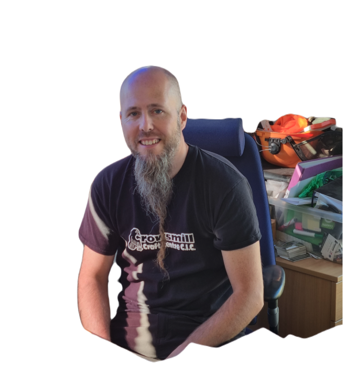 Man with a beard and long goatee sitting in a blue office chair, wearing a black t-shirt, with colorful storage boxes and backpacks in the background.
