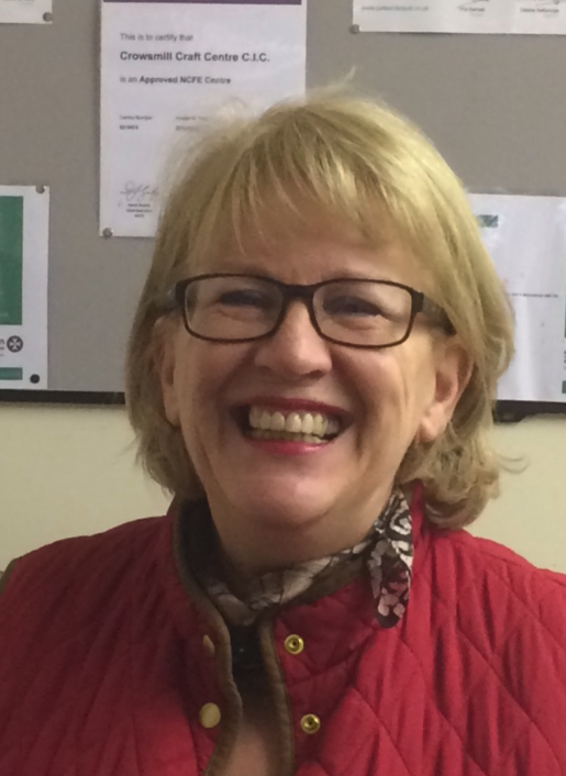 Smile of a woman with blonde hair, wearing glasses, a red quilted jacket, and a patterned scarf. Background shows a bulletin board with papers.