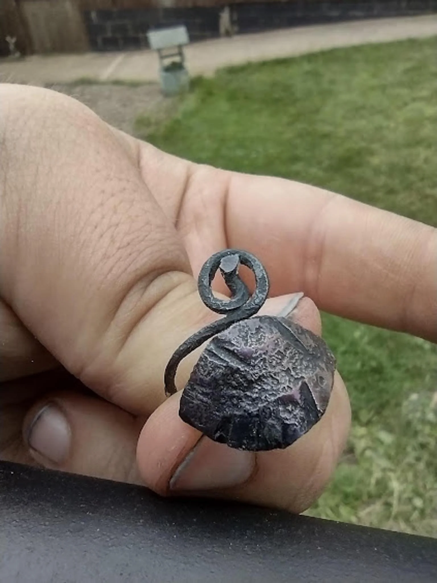 Close-up of a person's hand holding a small, dark, round, textured stone with a curled metal wire attached, outdoors with grass and a building in the background.