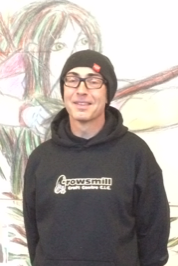 A young man wearing glasses, a black beanie, and a black hoodie with logo, standing in front of a colorful, childlike drawing of a girl with blonde hair and a bow.