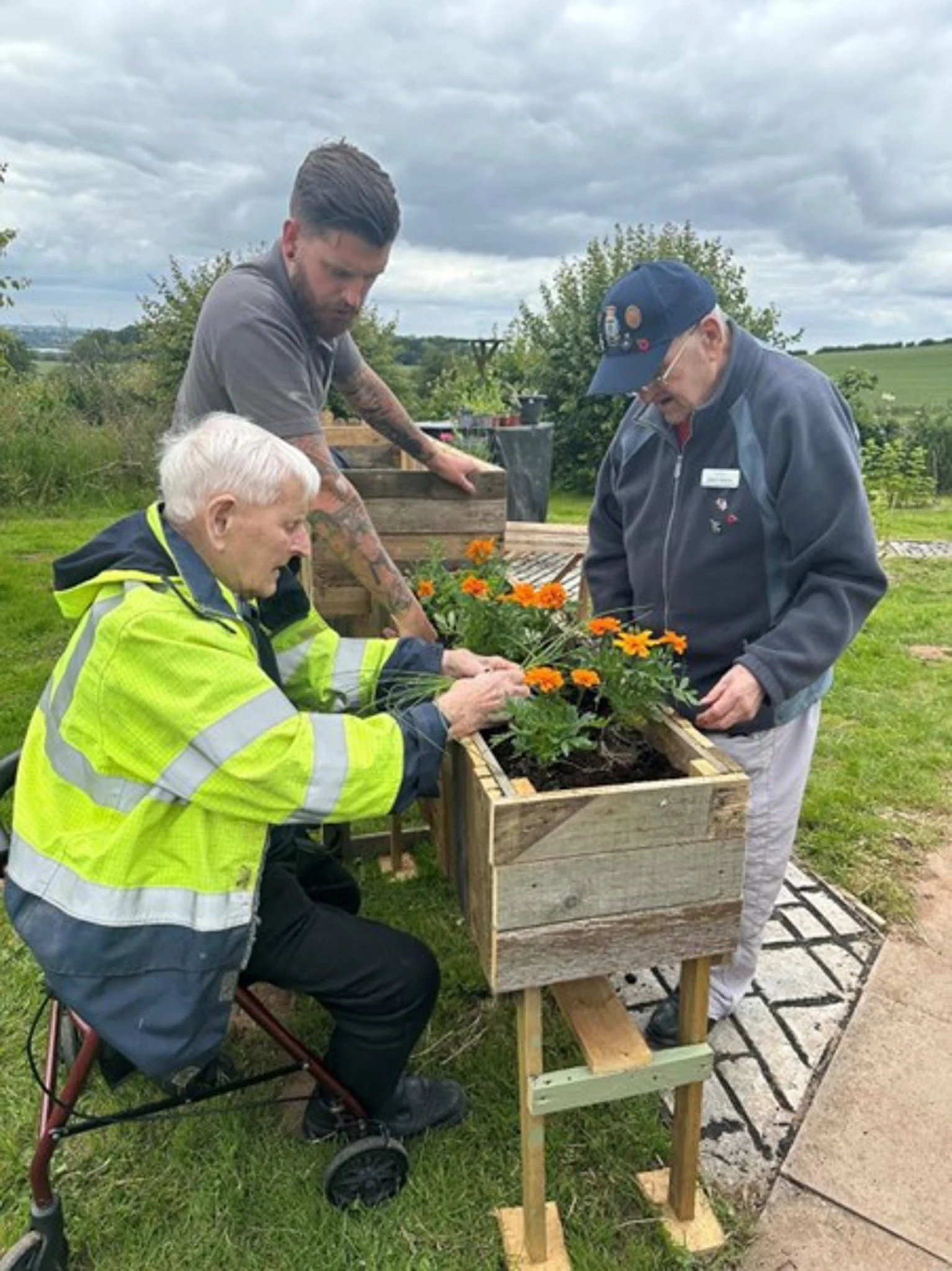 Three men are planting flowers in a wooden garden box outdoors, with green grass and trees in the background under a cloudy sky.