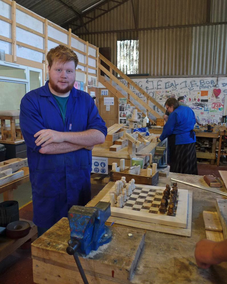 A young man with red hair and beard wearing a blue work uniform standing with arms crossed in a woodworking workshop, with other people working at tables, a chessboard, and wooden projects around.