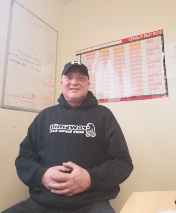 A man wearing a black cap and black hoodie with 'Cowshim' and 'Coach George C.L.C.' written on it, sitting indoors with a whiteboard and a large calendar behind him.