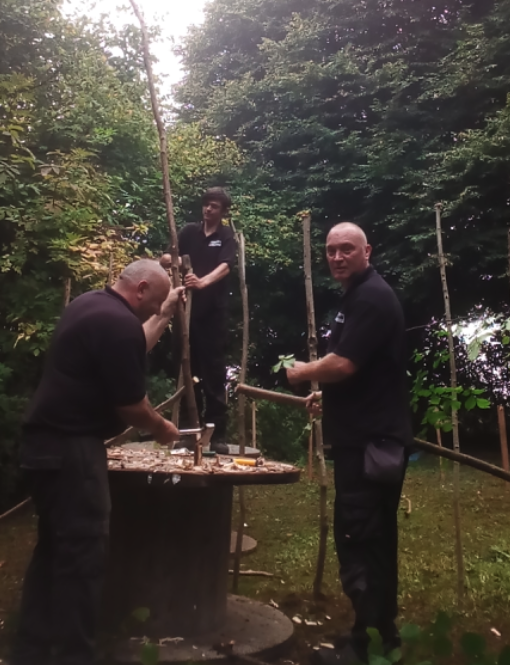 Three men working outdoors among trees, building a structure with wooden poles. One man is hammering a wooden piece onto a table, while another holds a pole, and a third is standing on a platform holding a pole vertically.