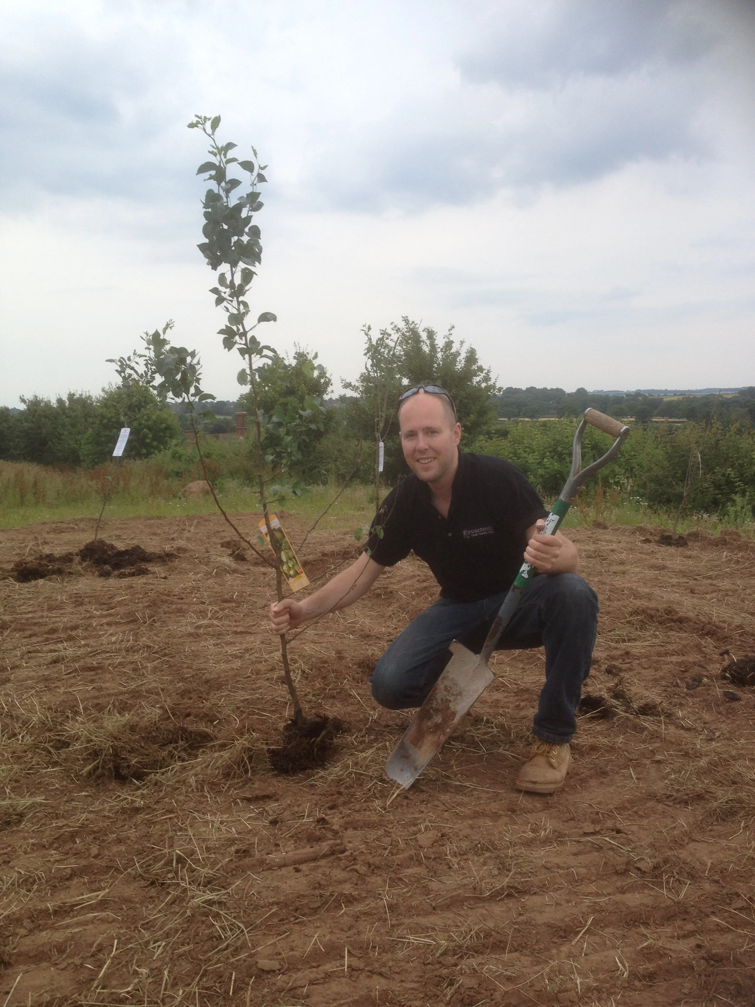 A man crouching in a field holding a small tree sapling and a shovel, with a cloudy sky overhead.