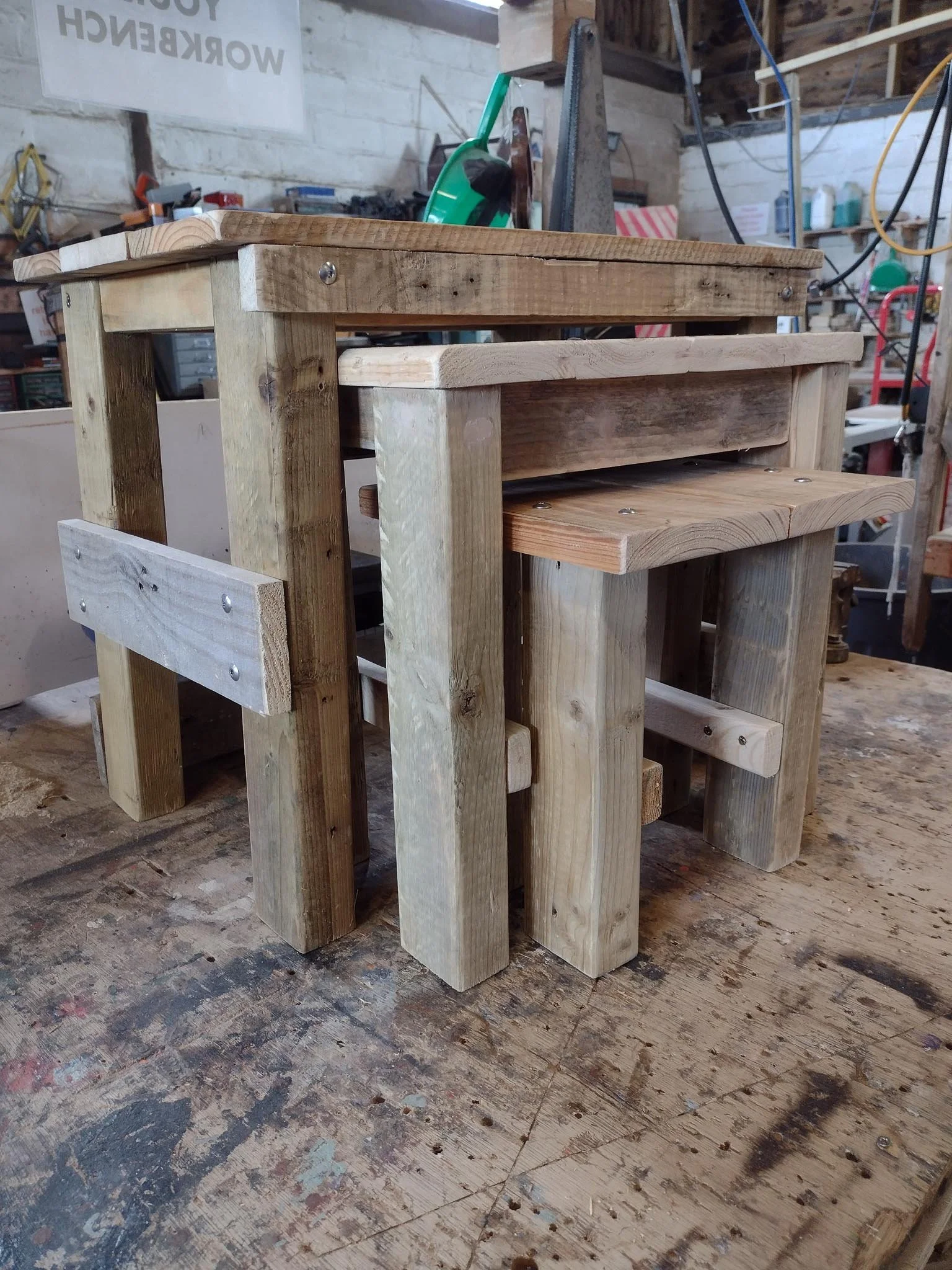 A small wooden stool made of unfinished wood planks with four legs, placed on a workbench in a woodworking shop.