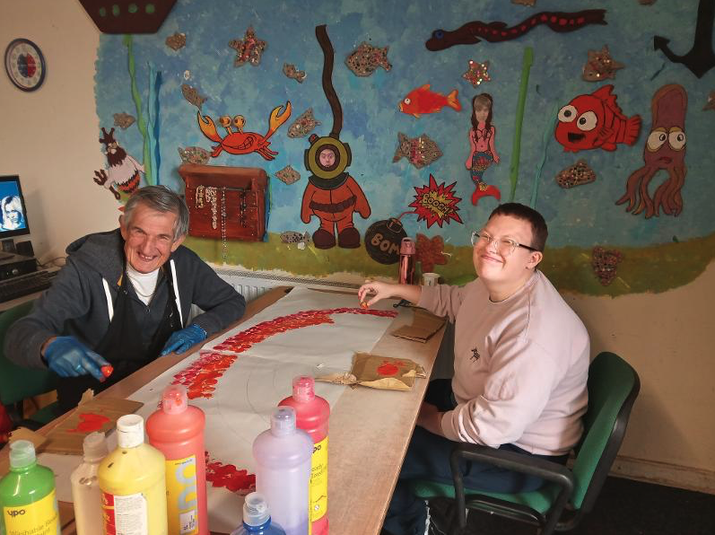Two smiling individuals working on a large Valentine’s Day paper craft project at a table, surrounded by colorful art supplies including bottles of paint. The background features a vibrant underwater-themed mural with cartoon-style sea creatures and characters.
