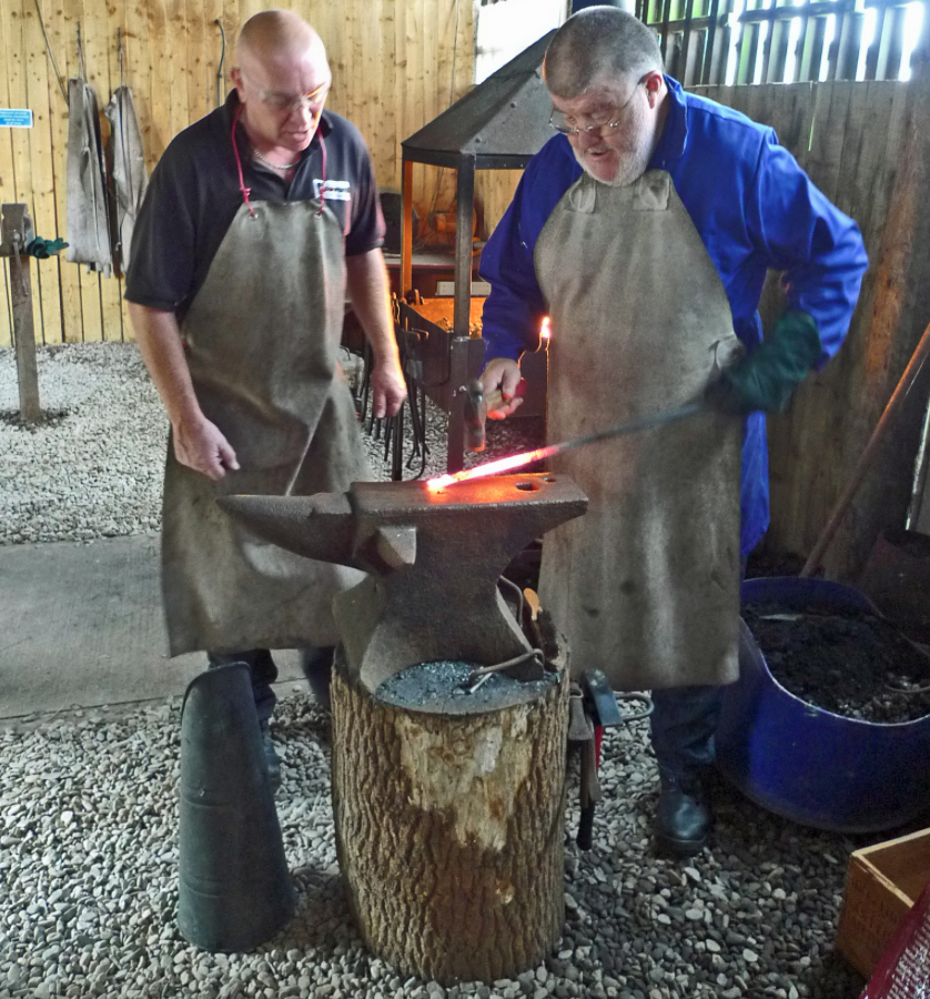 Two men practicing blacksmithing in a workshop. One is hammering hot metal on an anvil, while the other watches.