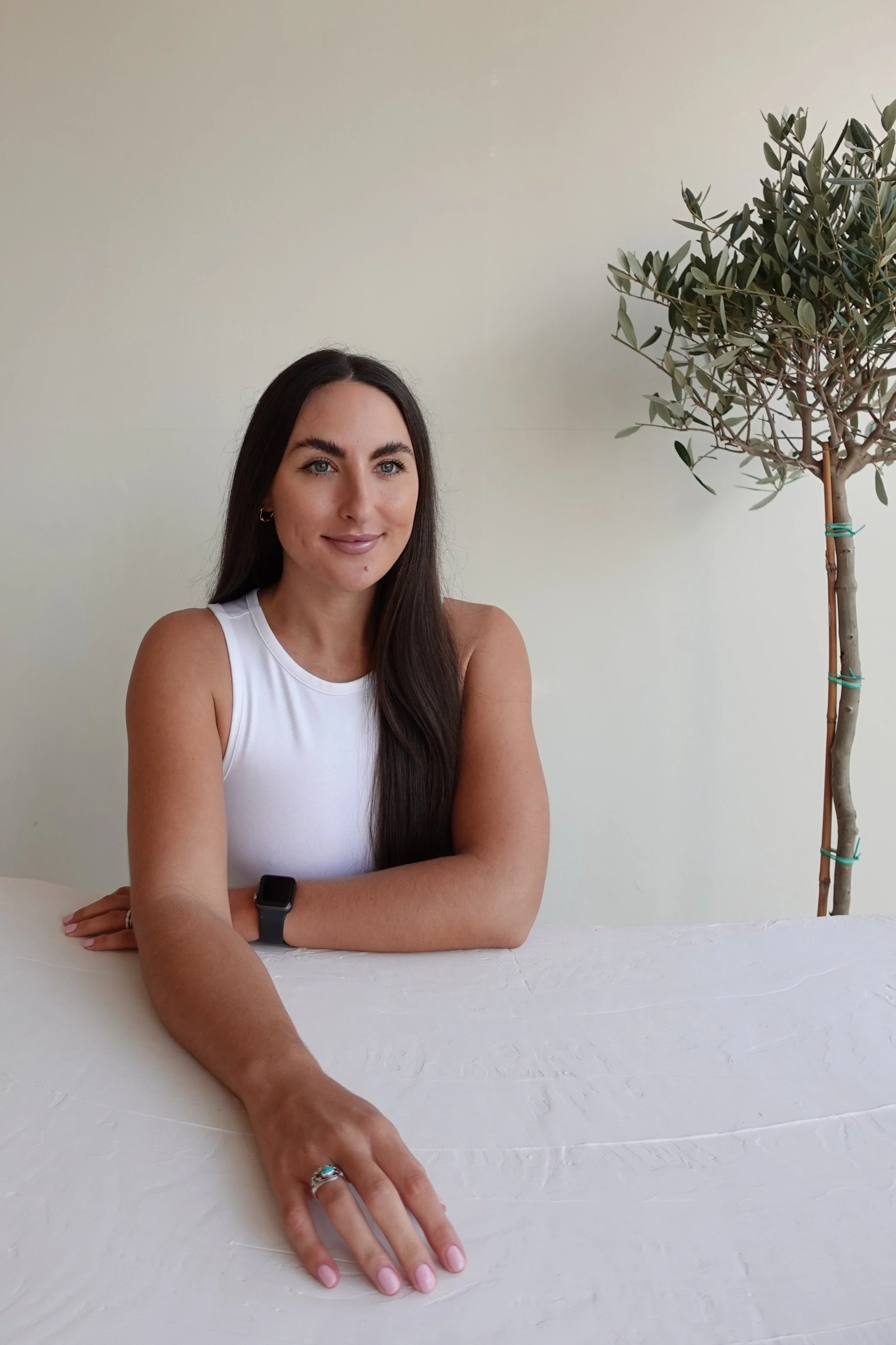 Woman with long dark hair sitting at a table wearing a white tank top and a smartwatch, near an olive tree.