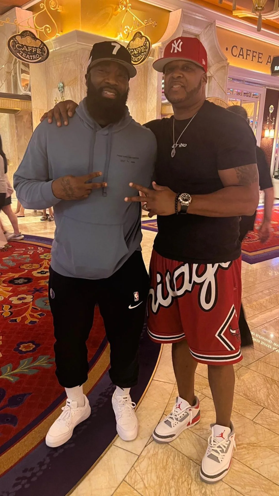Two men posing together in a lobby, both wearing basketball sneakers and casual clothing, with one in a gray hoodie and the other in a black t-shirt and red shorts, both making peace signs.