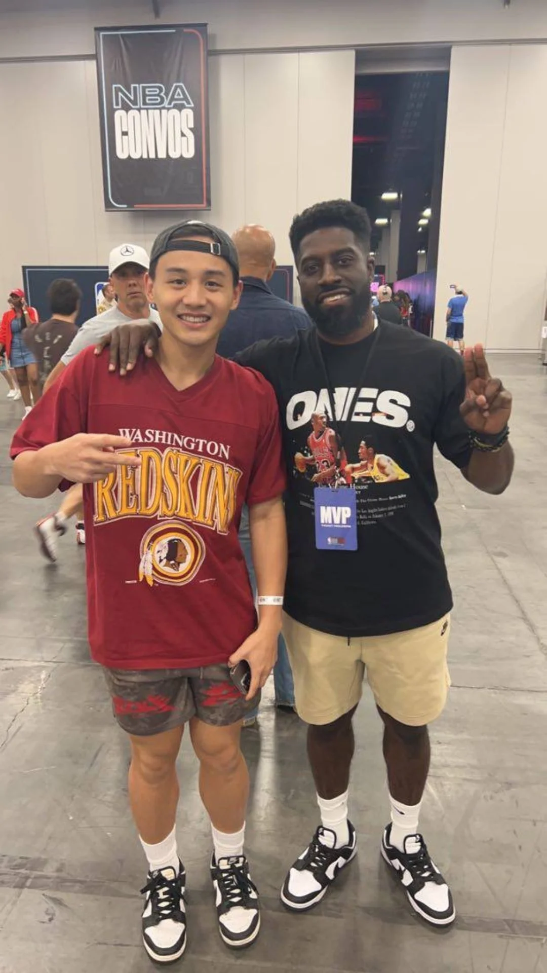 Two men posing inside an event hall, one wearing a Washington Redskins shirt and the other wearing a black T-shirt with a basketball graphic and MVP badge, both making hand gestures and smiling.